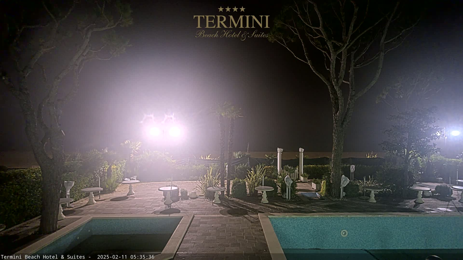 A nighttime scene shows a hazy outdoor patio area with a swimming pool, small tables, and trees, illuminated by bright lights.