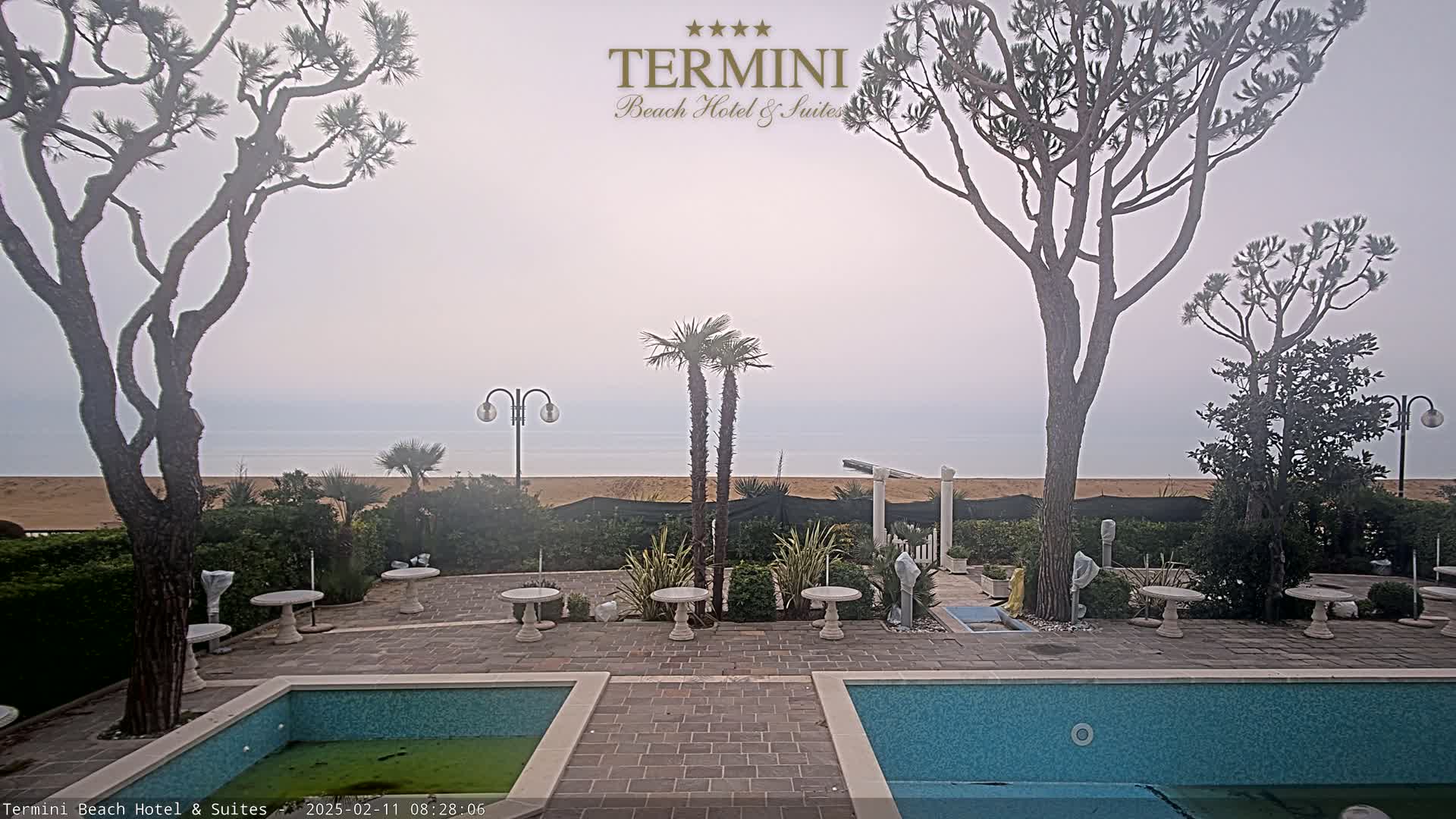 A hazy view of a calm ocean beach is seen from a courtyard with two pools and several small tables, framed by tall trees and landscaping.