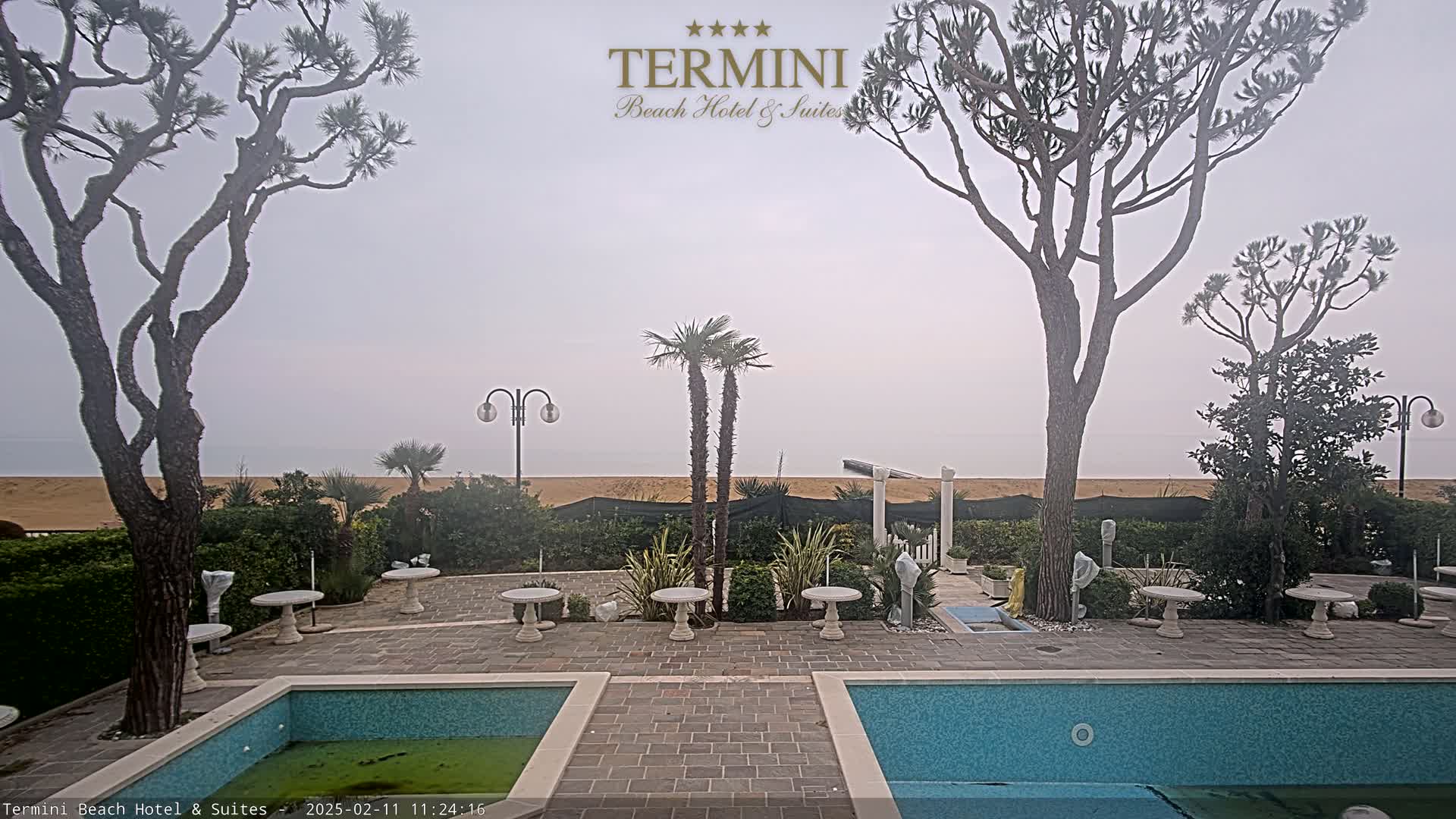 A hazy outdoor scene shows a patio area with two swimming pools, several small tables, and trees surrounding a sandy beach.