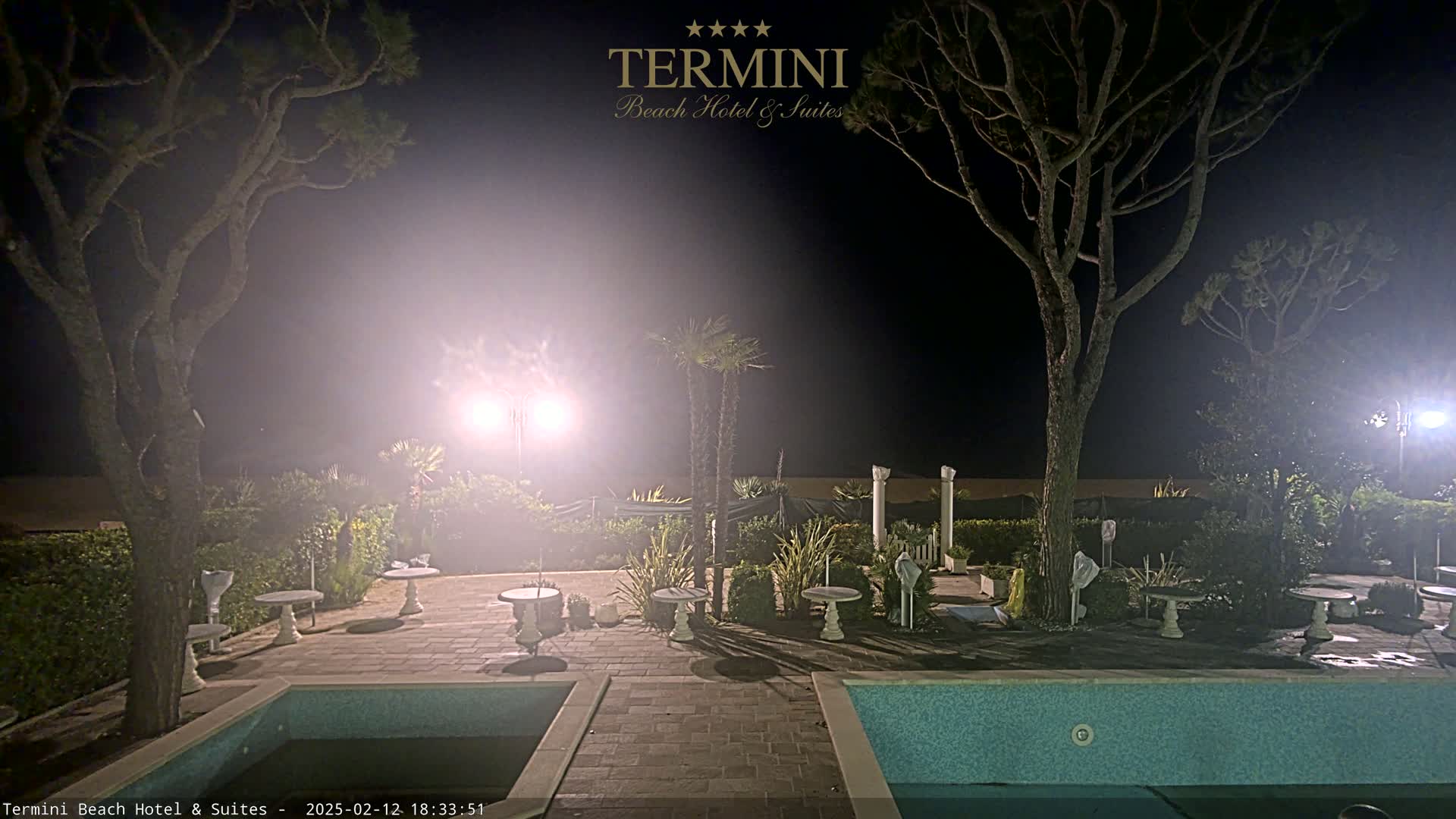 A nighttime scene shows a poolside patio area with several small tables and chairs surrounding a swimming pool, illuminated by bright lights under a hazy, dark sky.