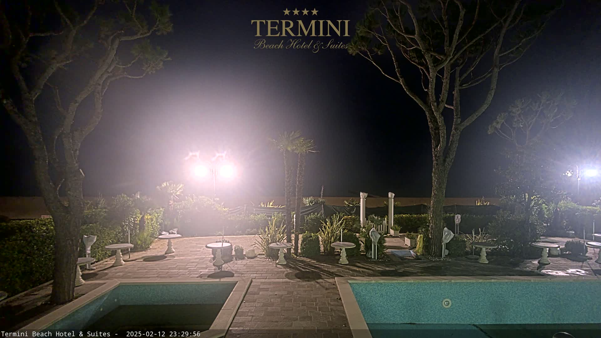 A hazy night scene shows a poolside patio area with several small tables and chairs, illuminated by bright lights, surrounded by trees and hedges, near a swimming pool.