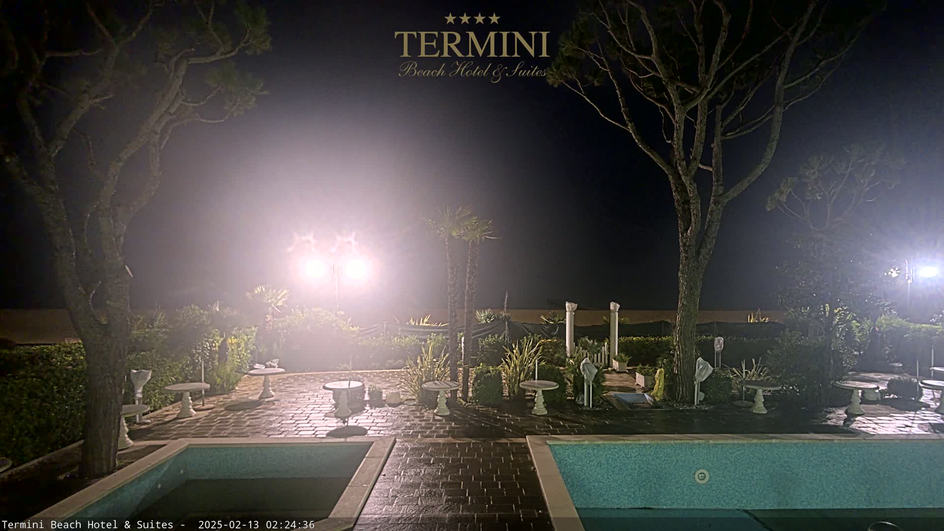 At night, under hazy, bright lights, an outdoor patio area with two pools is surrounded by trees and small tables.