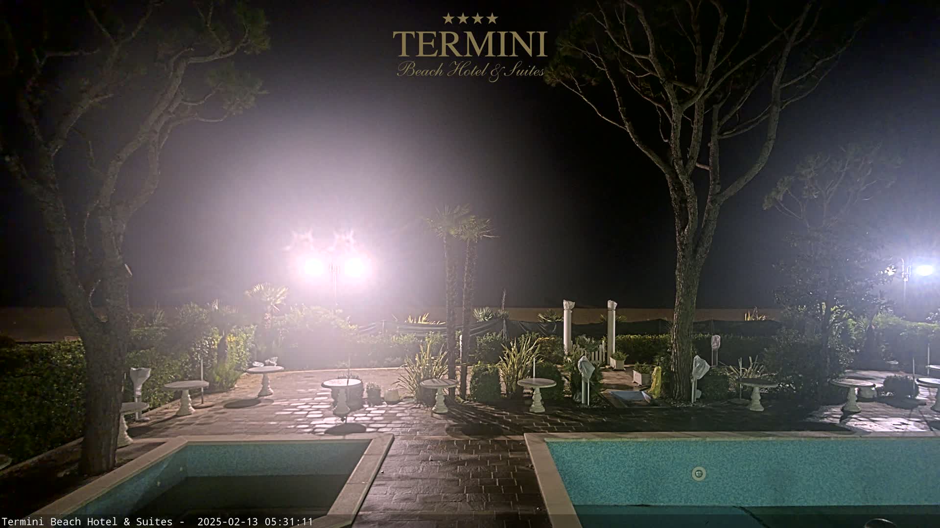At night, in foggy conditions, a patio area with two swimming pools and several small tables is illuminated by bright lights, surrounded by trees and landscaping.