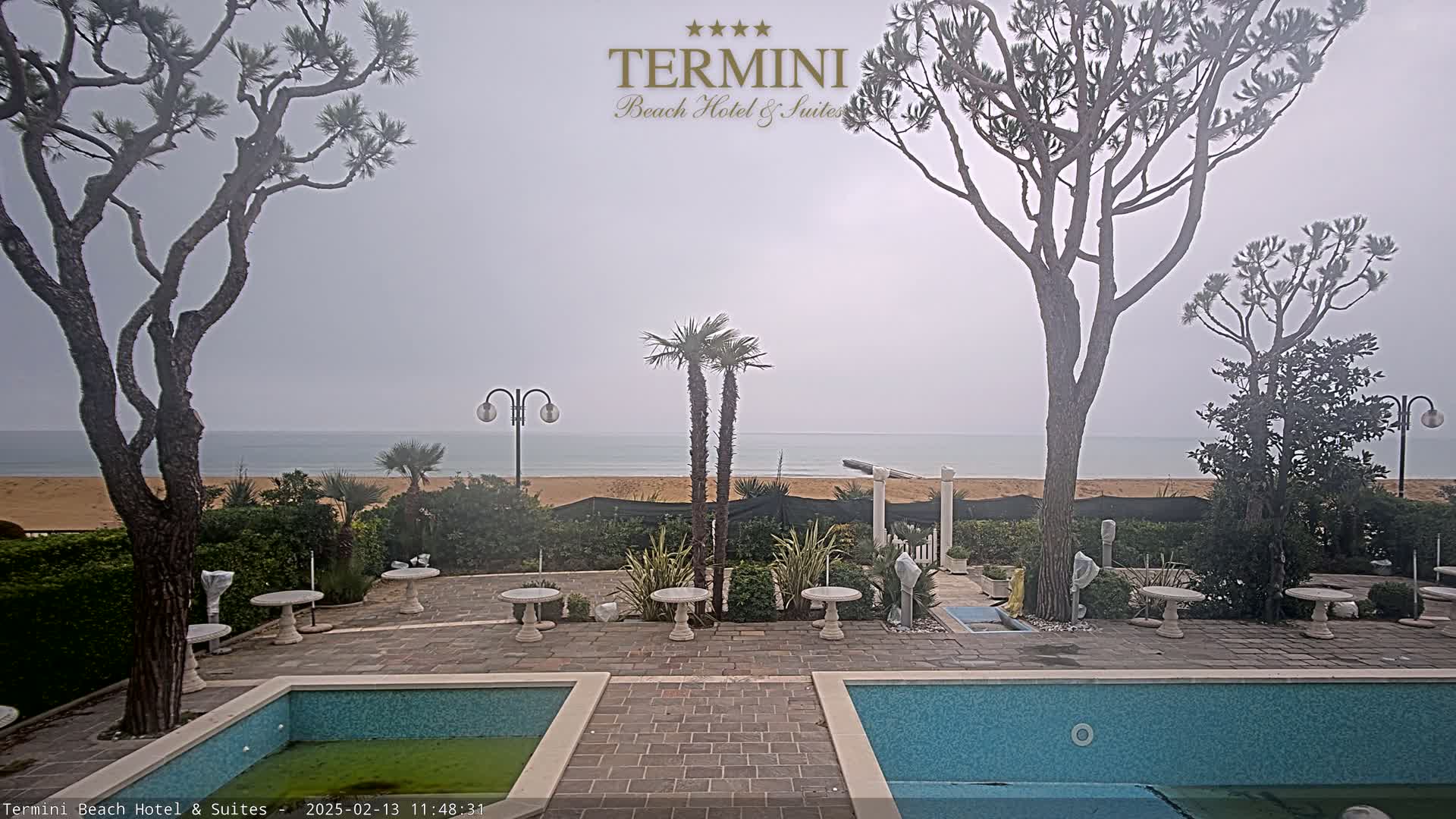 An overcast day reveals a view of a sandy beach and calm ocean from a patio area with two pools, several small tables, and palm and pine trees.