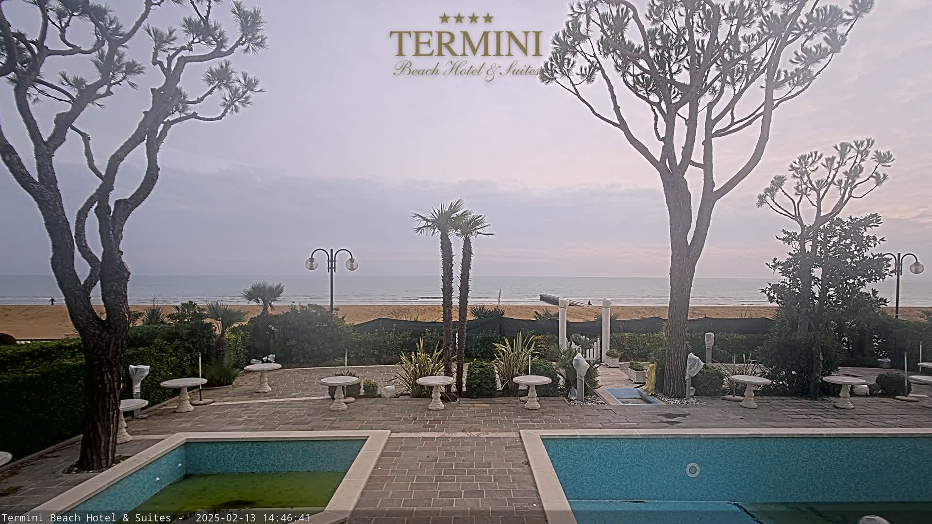 An outdoor patio area with two pools overlooks a calm ocean beach under an overcast sky.