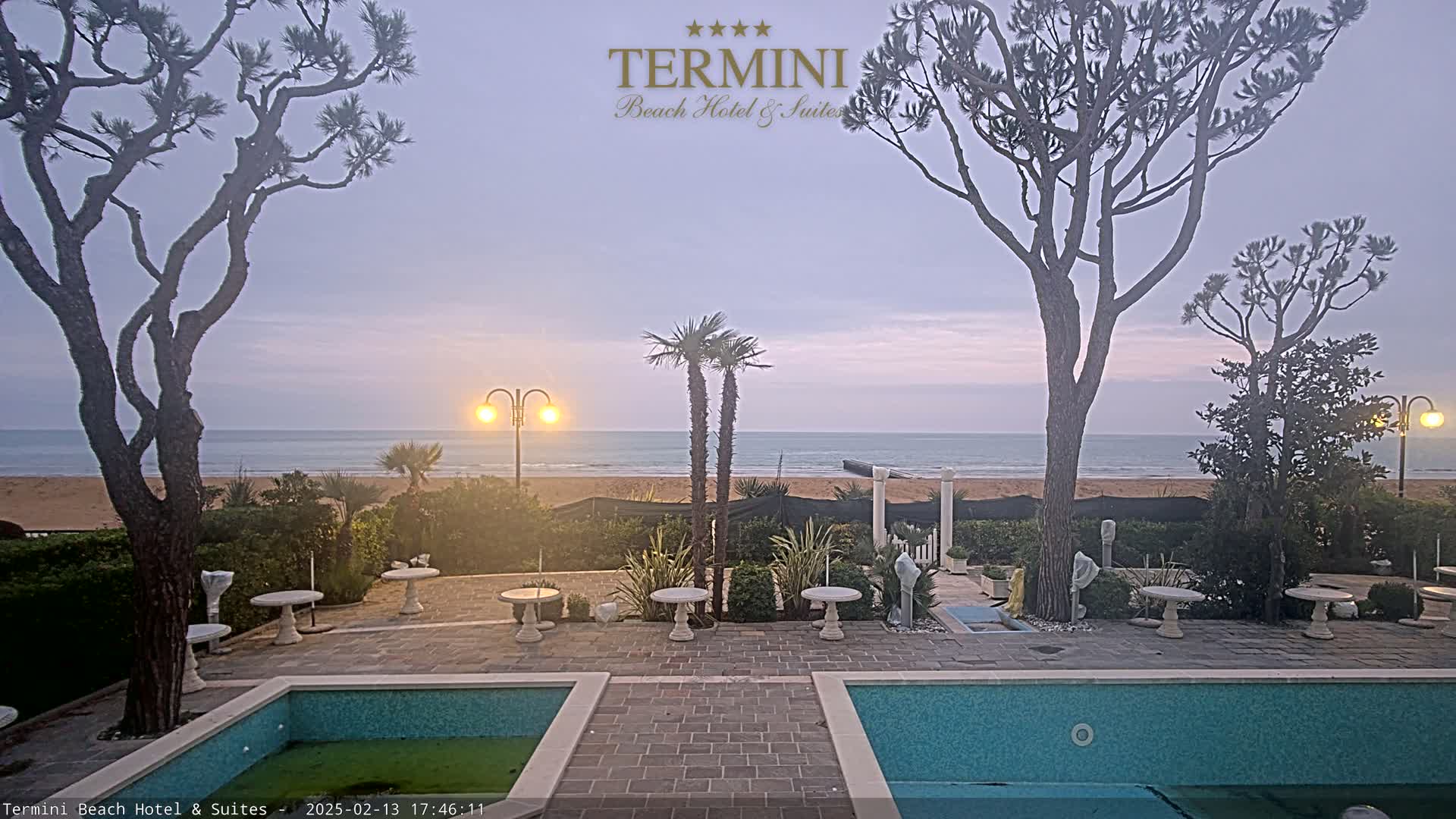 A tranquil outdoor scene shows a paved patio area with two pools and several small tables, flanked by trees and overlooking a calm ocean at sunset.