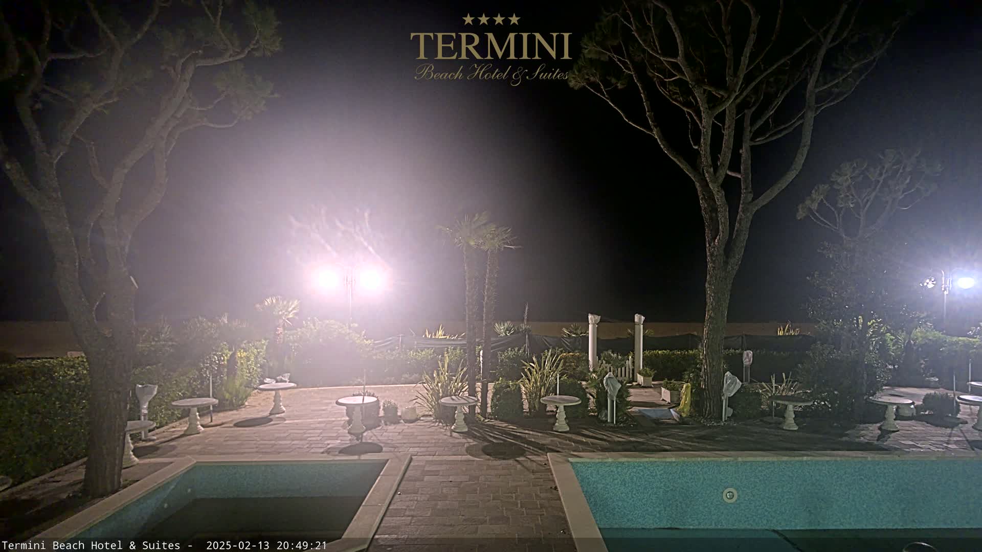 At night, under hazy conditions, a pool area with several small tables and trees is illuminated by bright lights.