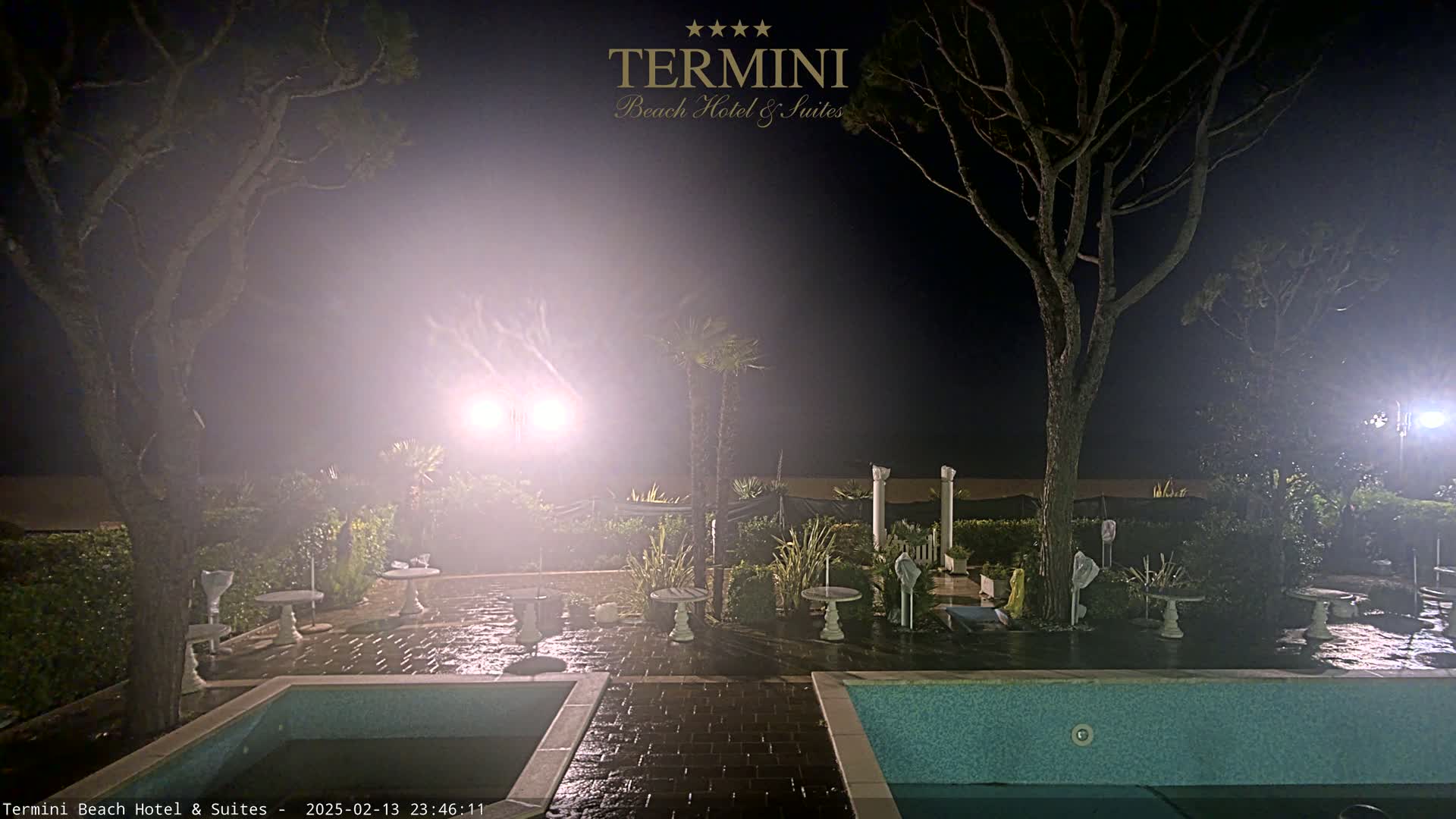 A hazy night scene shows an outdoor patio area with a swimming pool, several small tables and chairs, and trees, illuminated by bright lights.