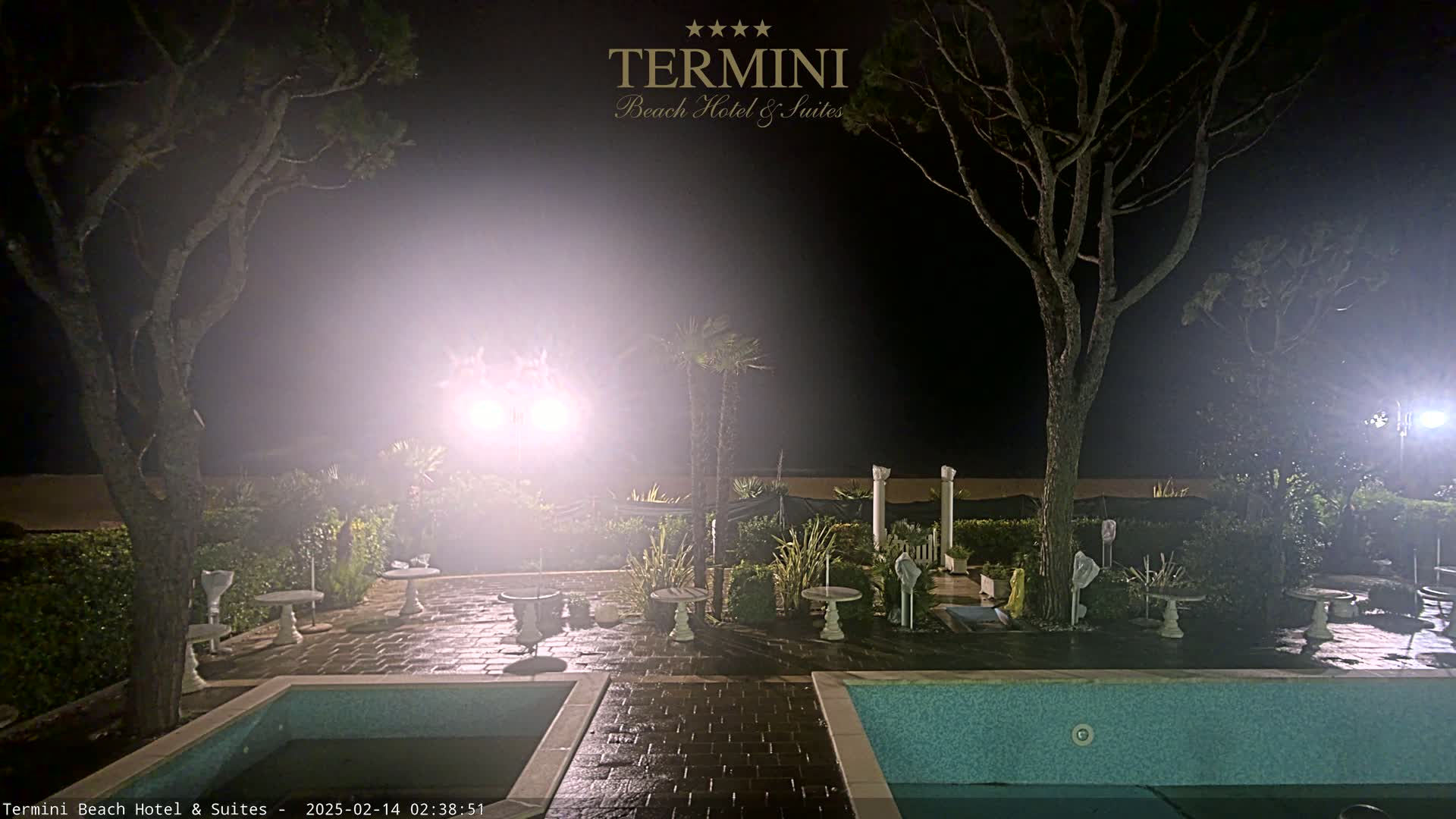 At night, a hazy light illuminates an outdoor patio area with two pools, small tables, and trees.