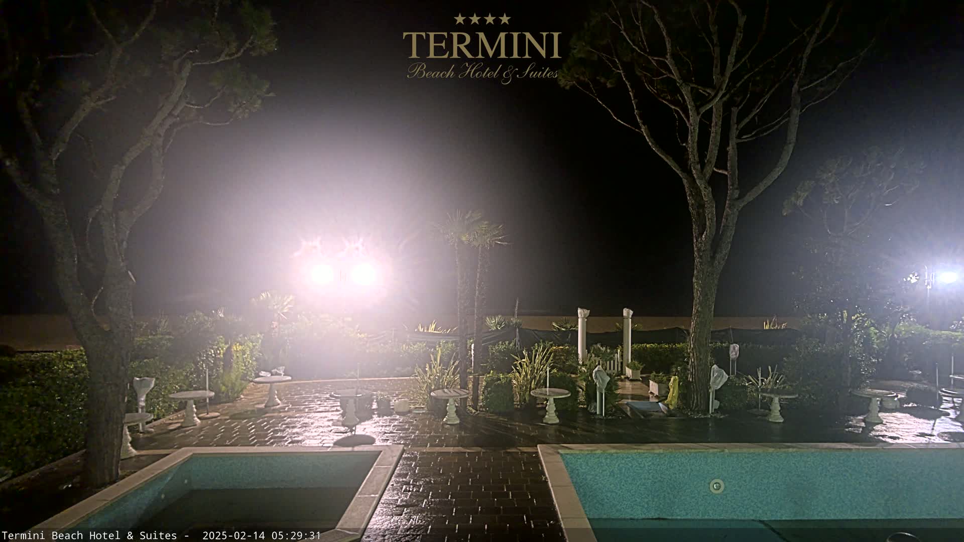 A nighttime view of a patio area with two swimming pools, surrounded by trees and small tables, under hazy, brightly lit conditions.