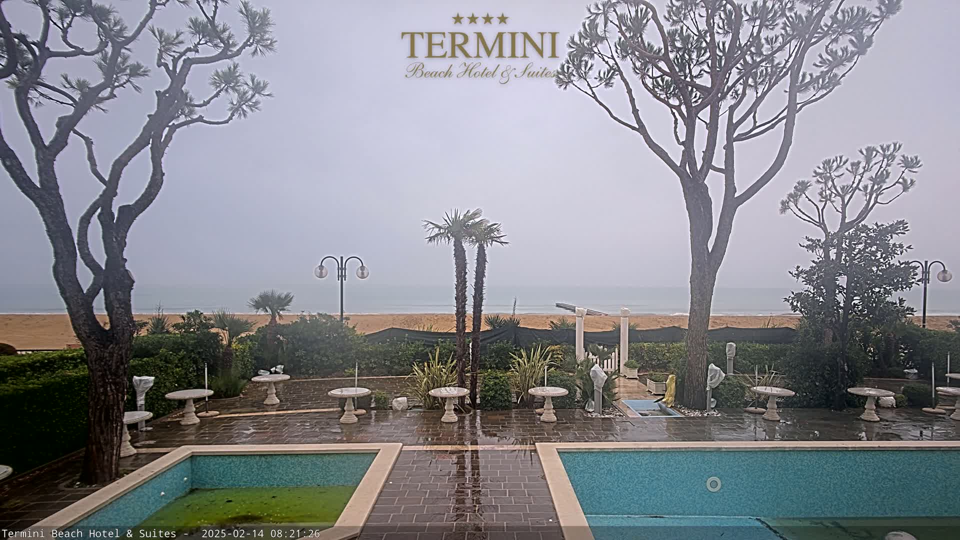 A misty seaside patio scene shows two pools, several small tables and chairs, and tall trees surrounding a sandy beach.