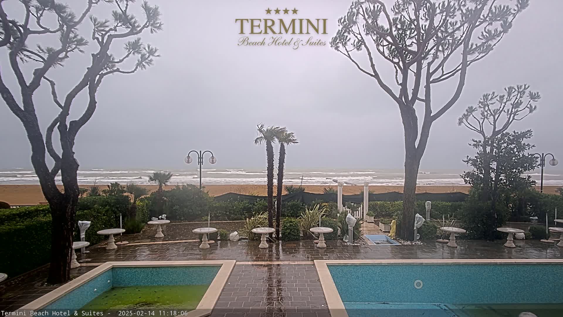 A wet patio with two pools overlooks a beach and stormy ocean under an overcast sky.