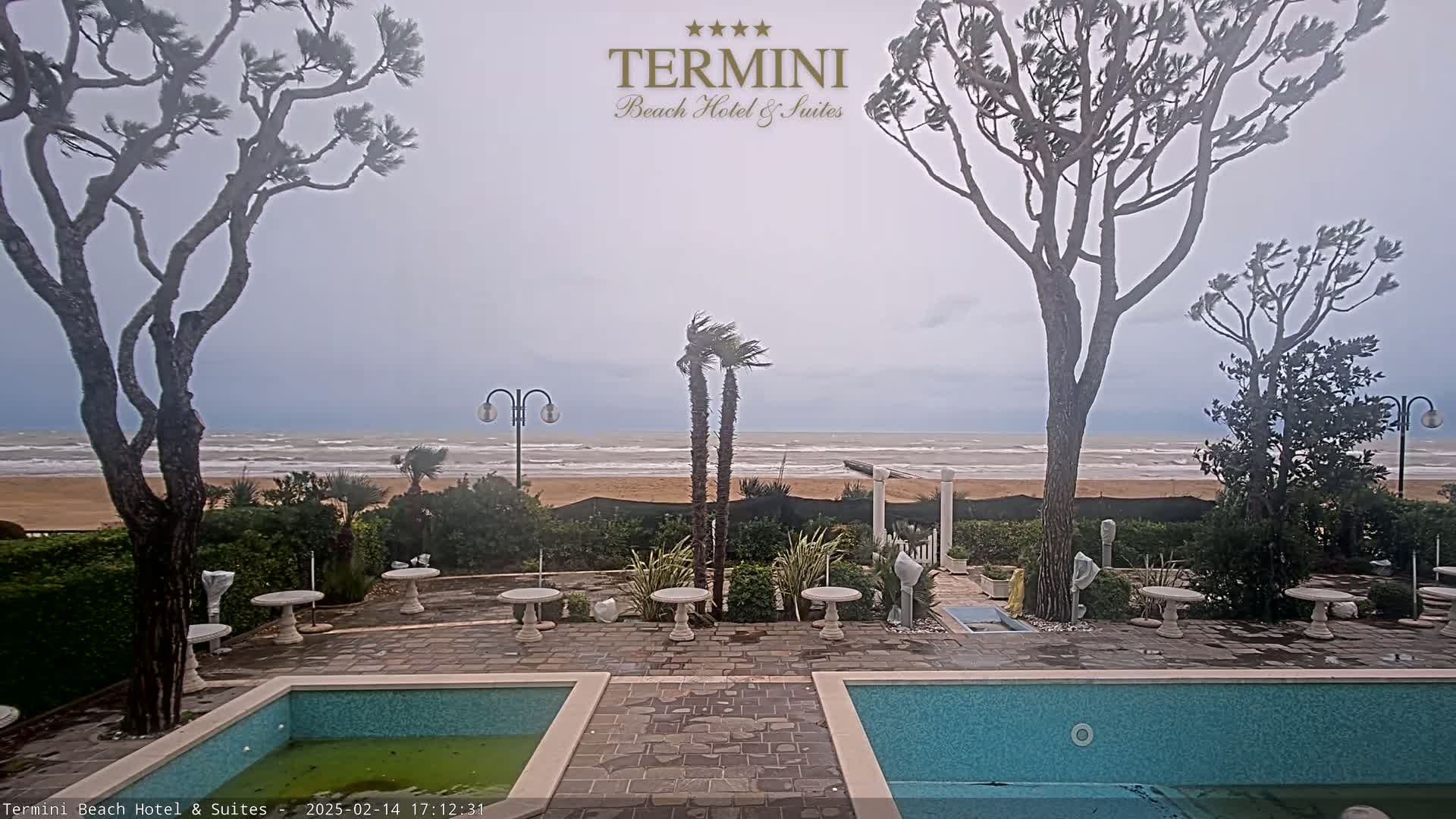 A windy, overcast day at a beachfront hotel reveals a patio with two swimming pools and several small tables, all situated between the building and a sandy beach with a choppy sea.