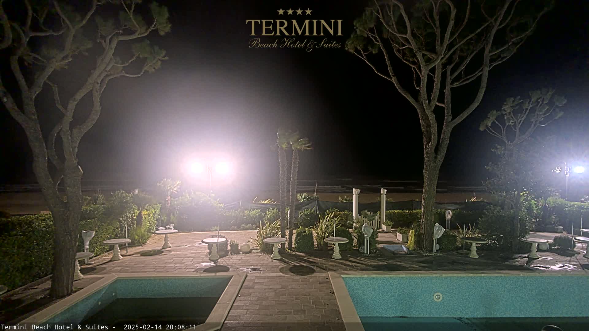 At night, a hazy beach is visible beyond a patio area with small tables, two swimming pools, and tall trees illuminated by bright lights.