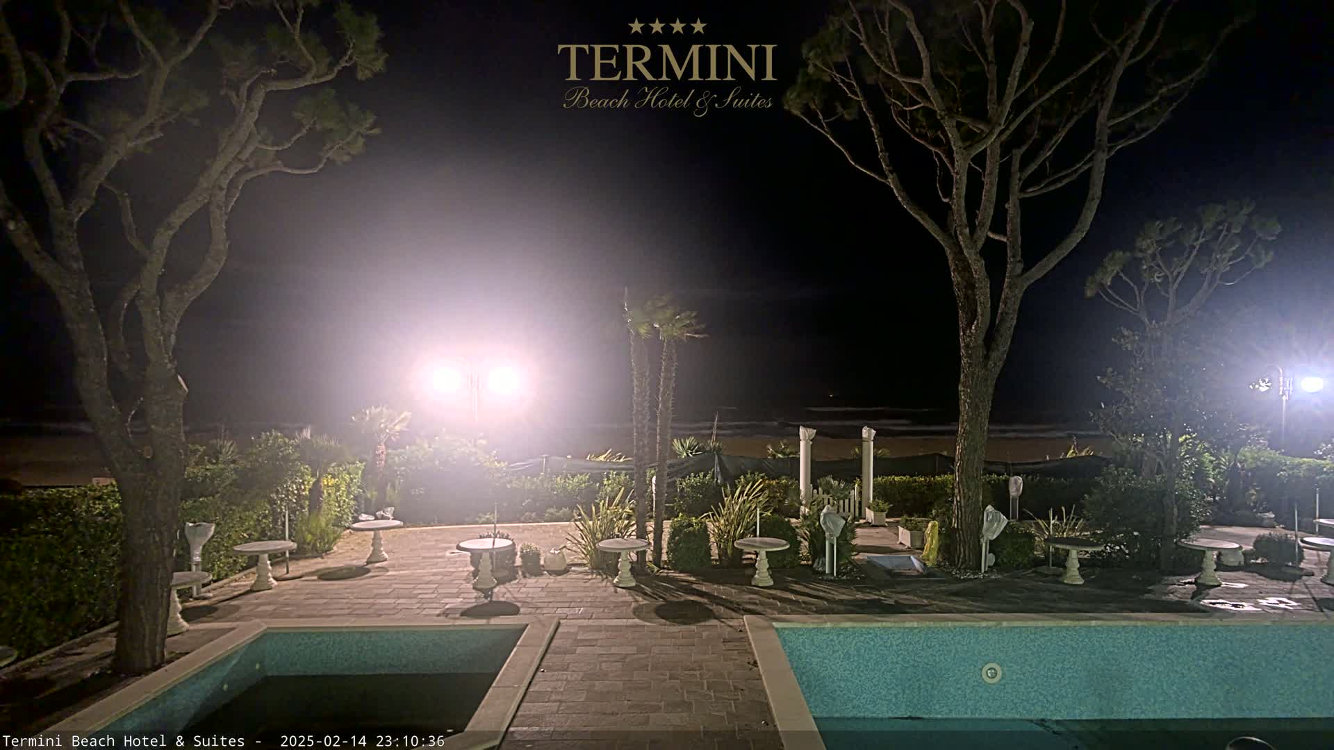At night, a resort patio with several small tables and a swimming pool overlooks a dimly lit beach under hazy conditions.