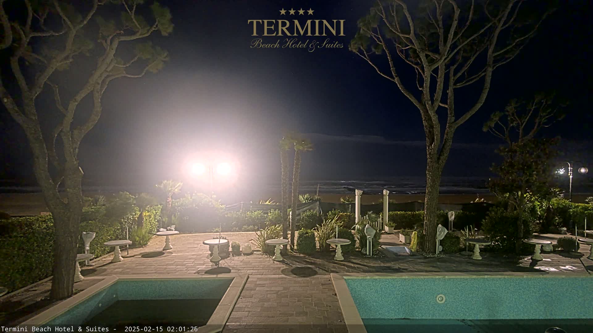 A nighttime view of a resort area shows a swimming pool, patio furniture, and trees surrounding a sandy beach under a dark, clear sky.