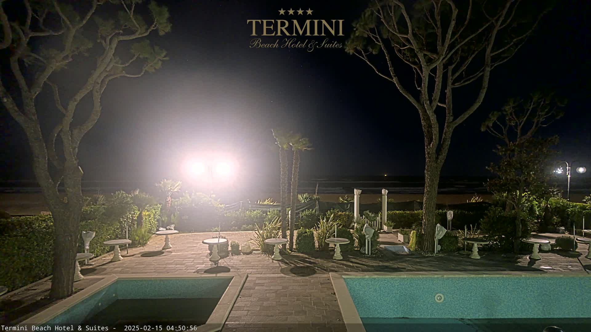 A nighttime view of a resort patio with a swimming pool, sparsely decorated with small tables and trees, overlooking a dark beach under a clear, starry sky.