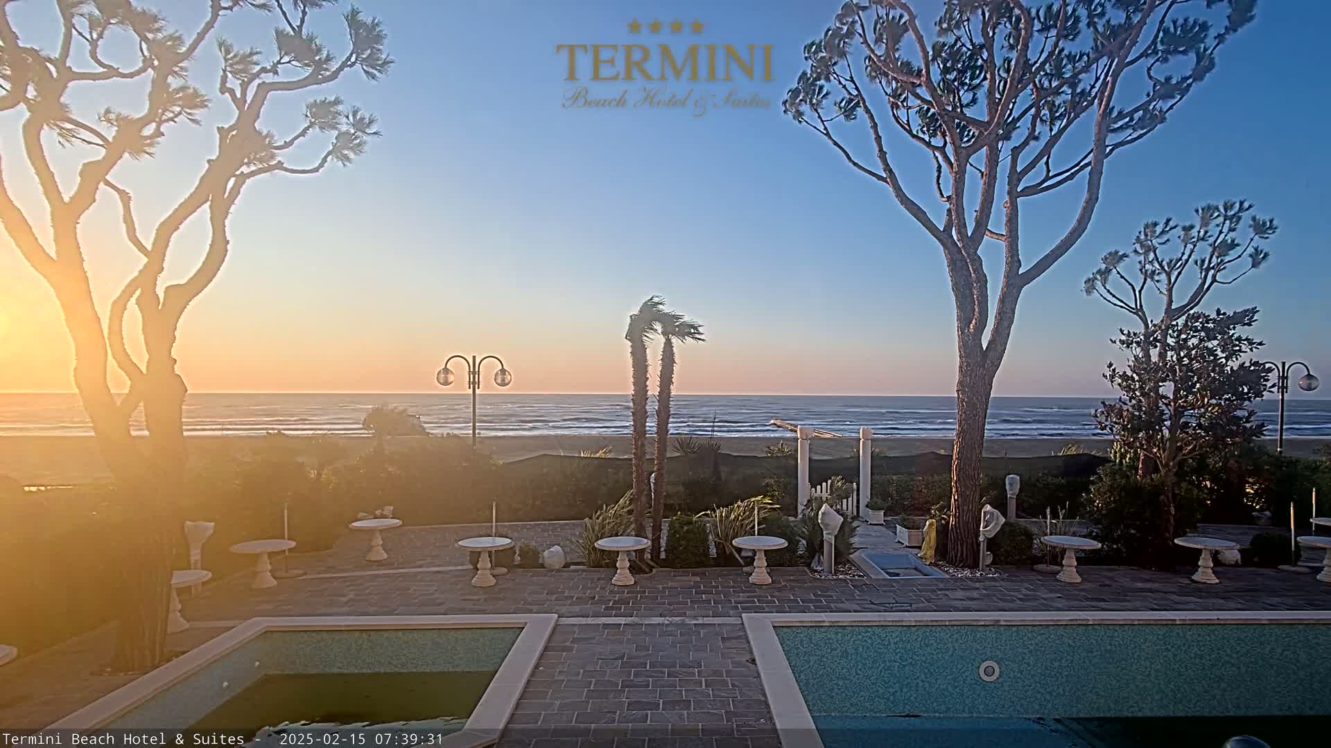 A sunrise over a calm ocean is viewed from a patio area with small tables, landscaping, and two pools.