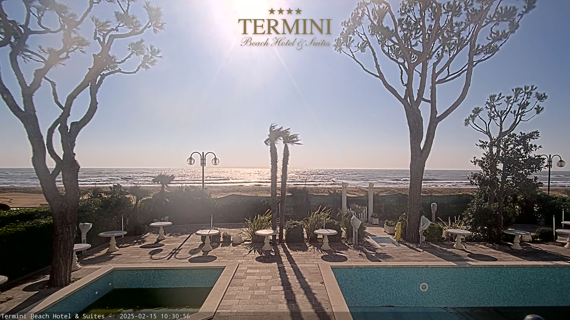 A sunny outdoor scene shows a patio area with two pools, small tables and chairs, and several trees overlooking a sandy beach and ocean with gentle waves.