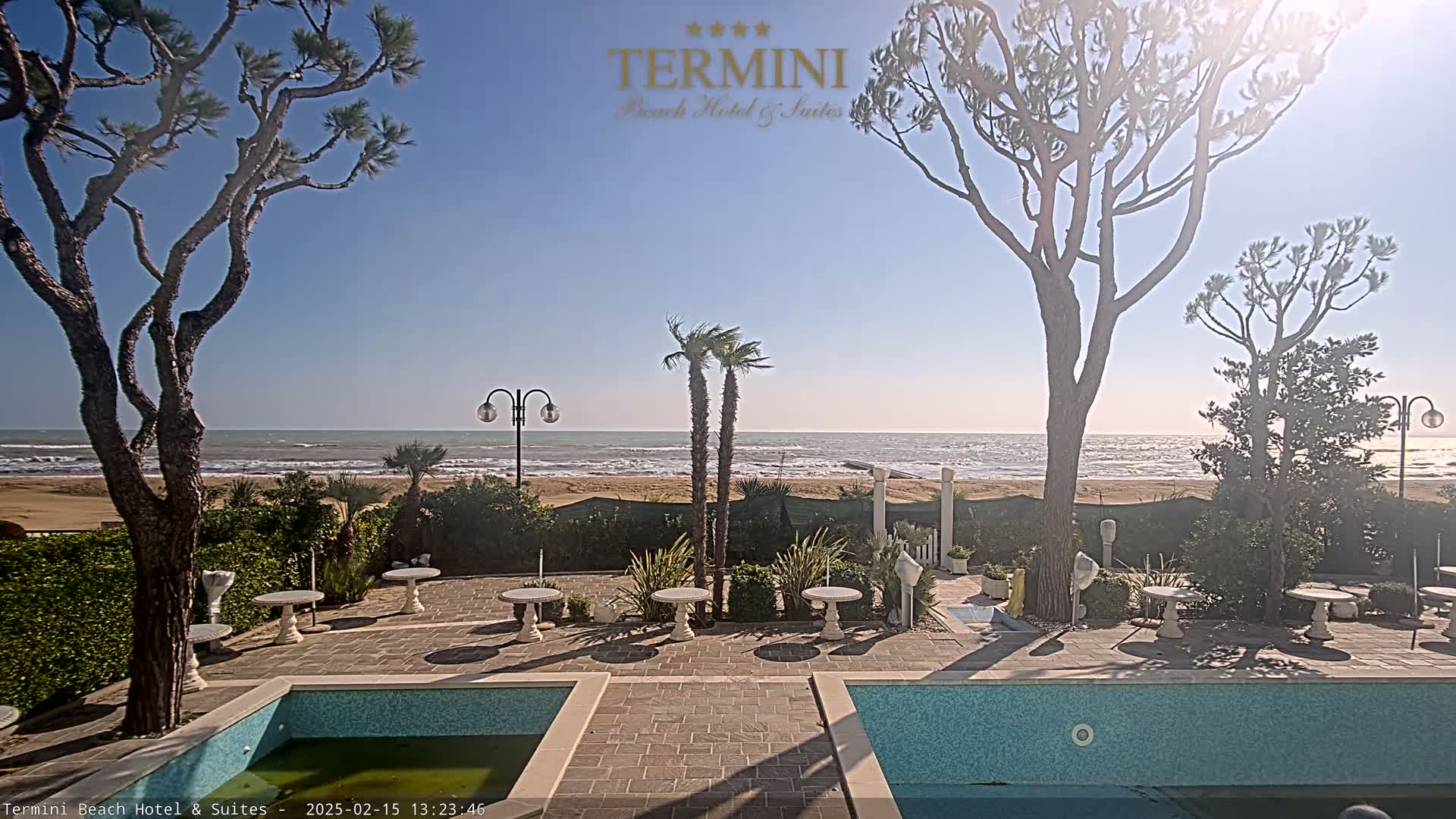 A patio with a pool overlooks a sandy beach and a calm ocean under a clear, sunny sky.