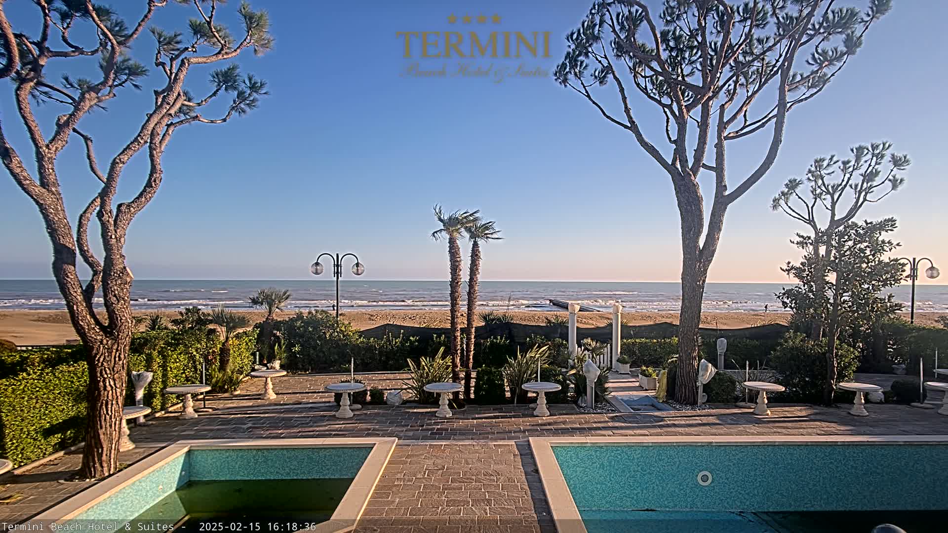 A patio area with two pools overlooks a sandy beach and calm ocean under a clear blue sky.