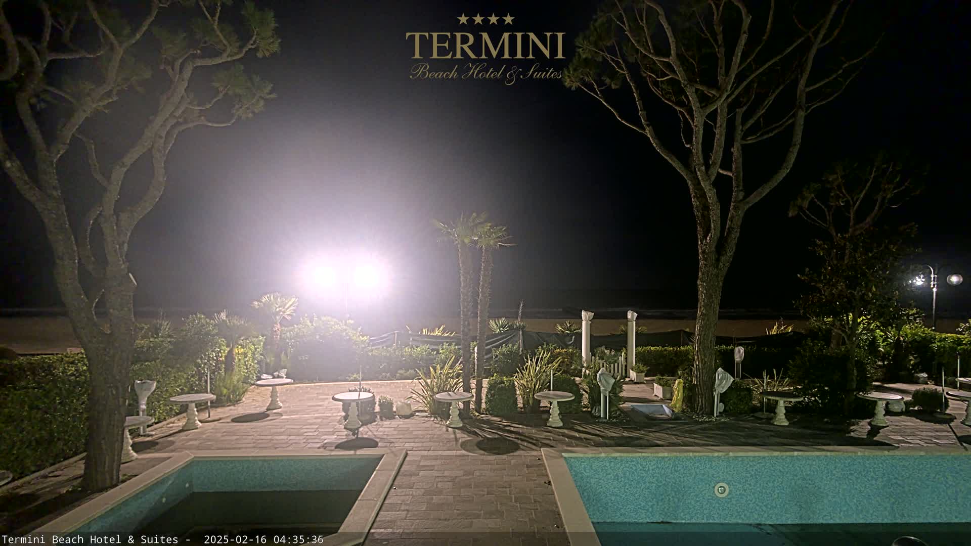 A nighttime view of a resort pool area with palm trees, small tables, and a beach visible in the background under a hazy sky.
