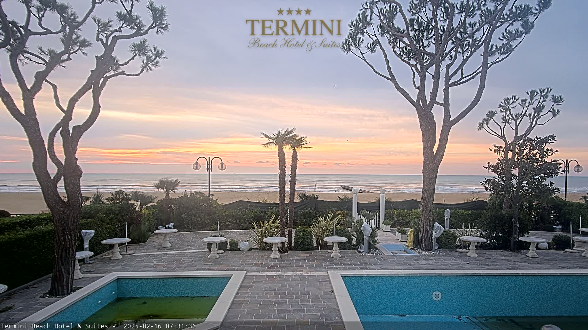 A tranquil sunrise over a calm ocean is visible from a patio area with two small pools and sparse landscaping.