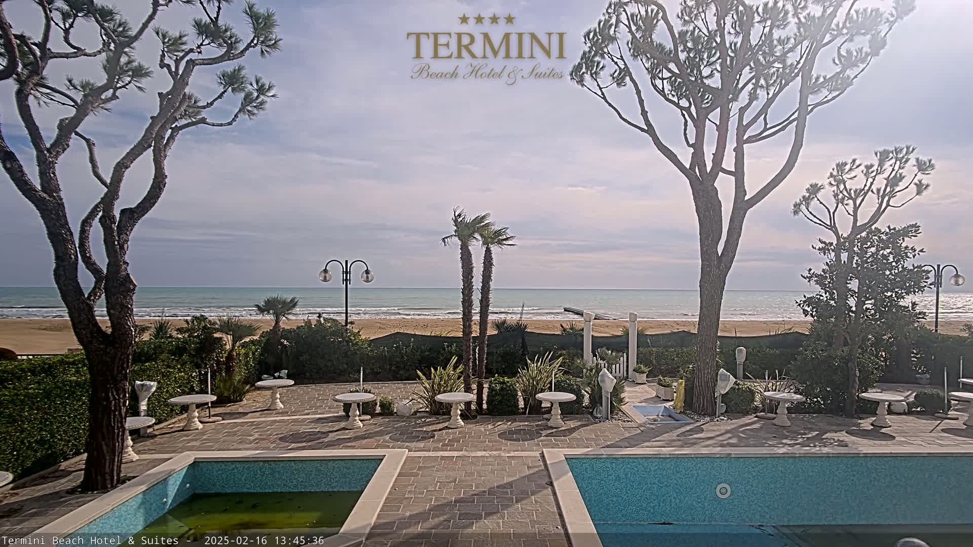 A tranquil beach scene is visible from a patio with two pools and small tables, under a partly cloudy sky.