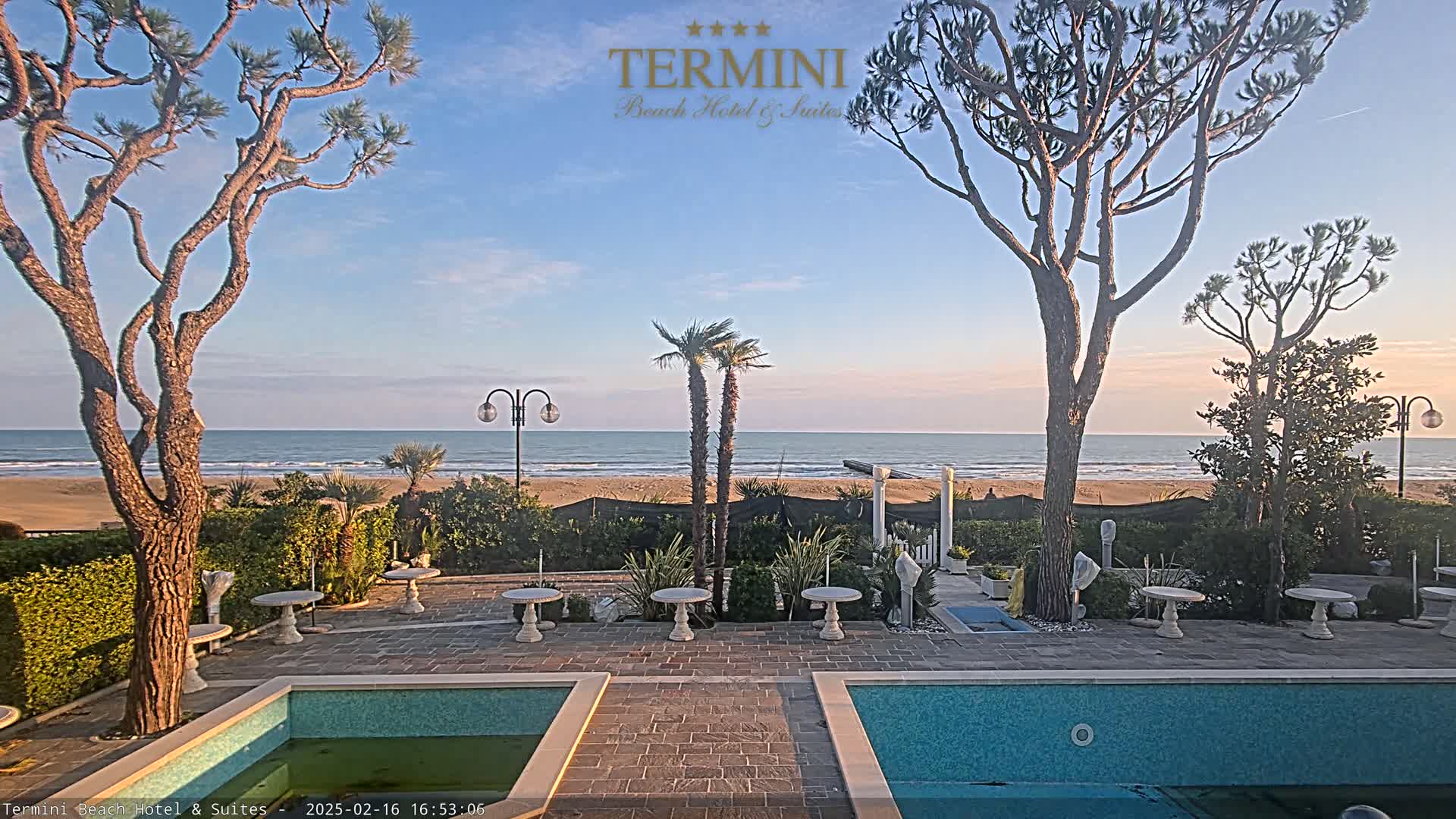 A courtyard with two pools overlooks a calm ocean beach under a clear sky.