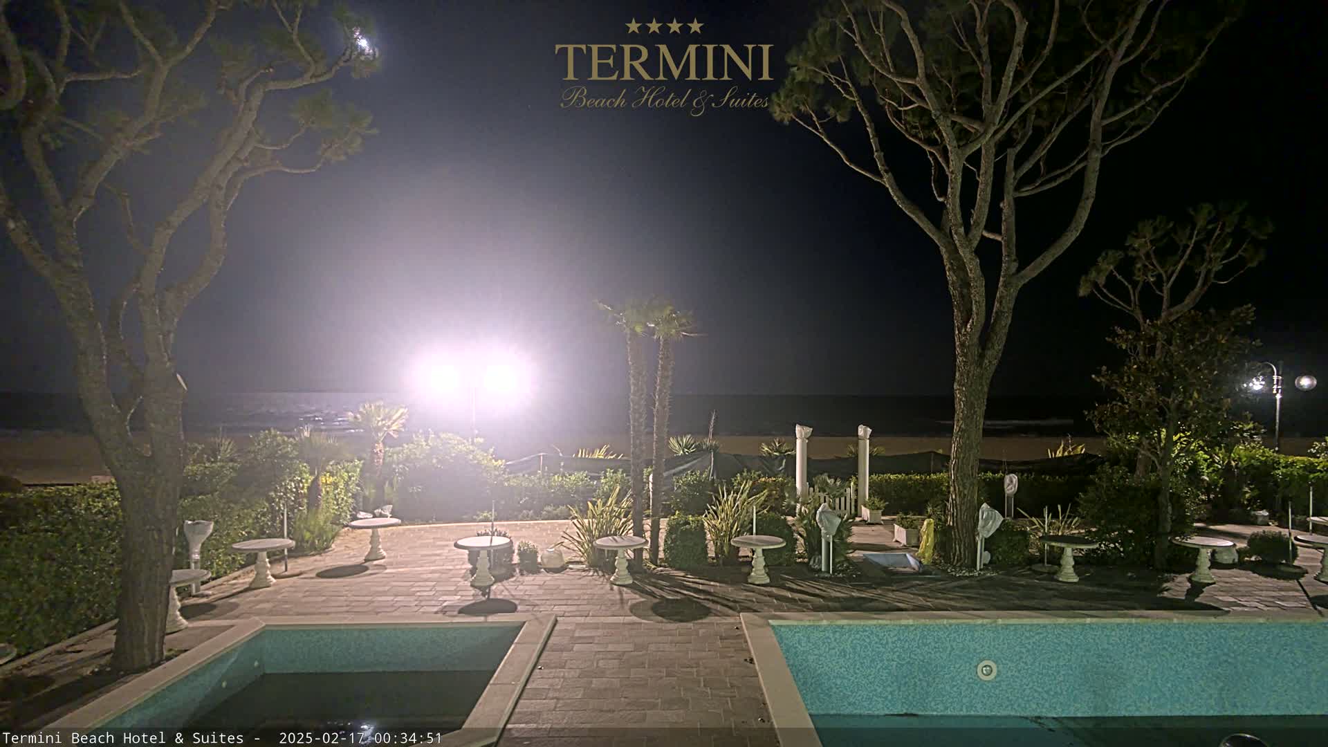A nighttime view of a resort pool area with palm trees, outdoor seating, and a calm ocean in the background under a mostly clear sky.