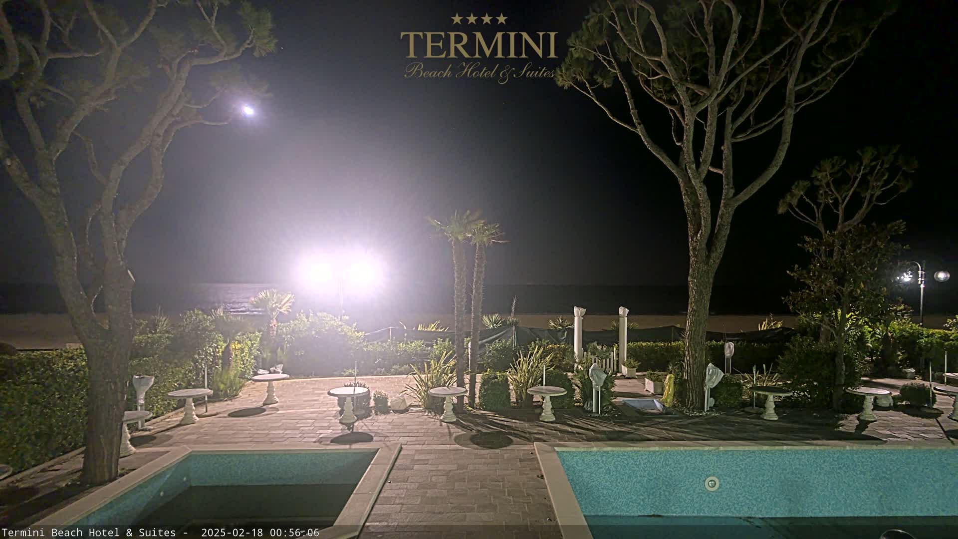 A nighttime view of a resort area with a swimming pool, patio furniture, and palm trees overlooking a calm ocean under a partly cloudy sky.