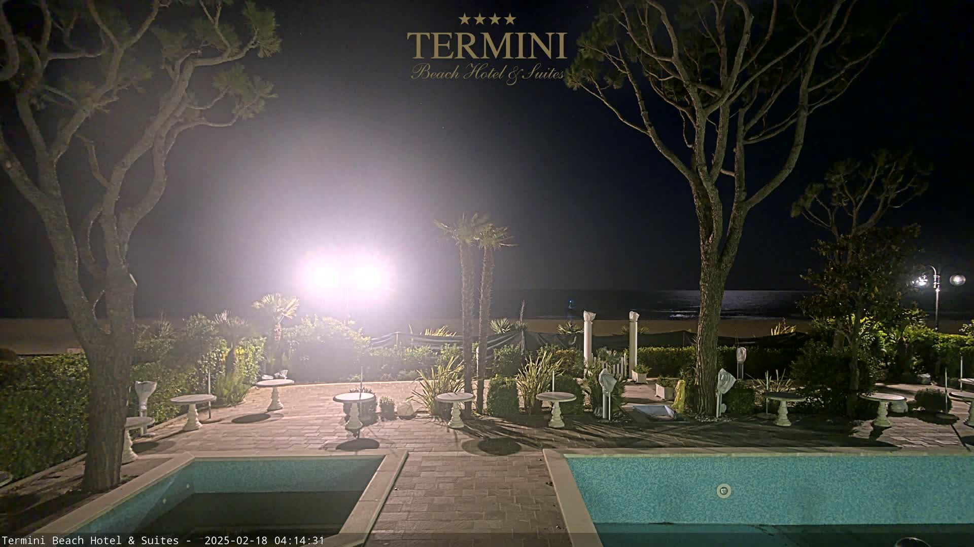 A nighttime view of a resort pool area with palm trees and a calm ocean visible in the distance under a mostly clear sky.