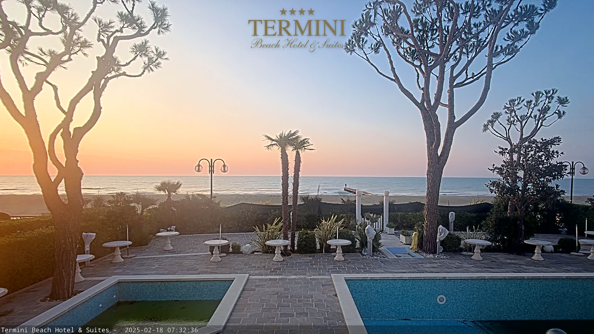A calm ocean at sunrise is visible beyond a patio area with two pools and several small tables, all surrounded by trees and landscaping.