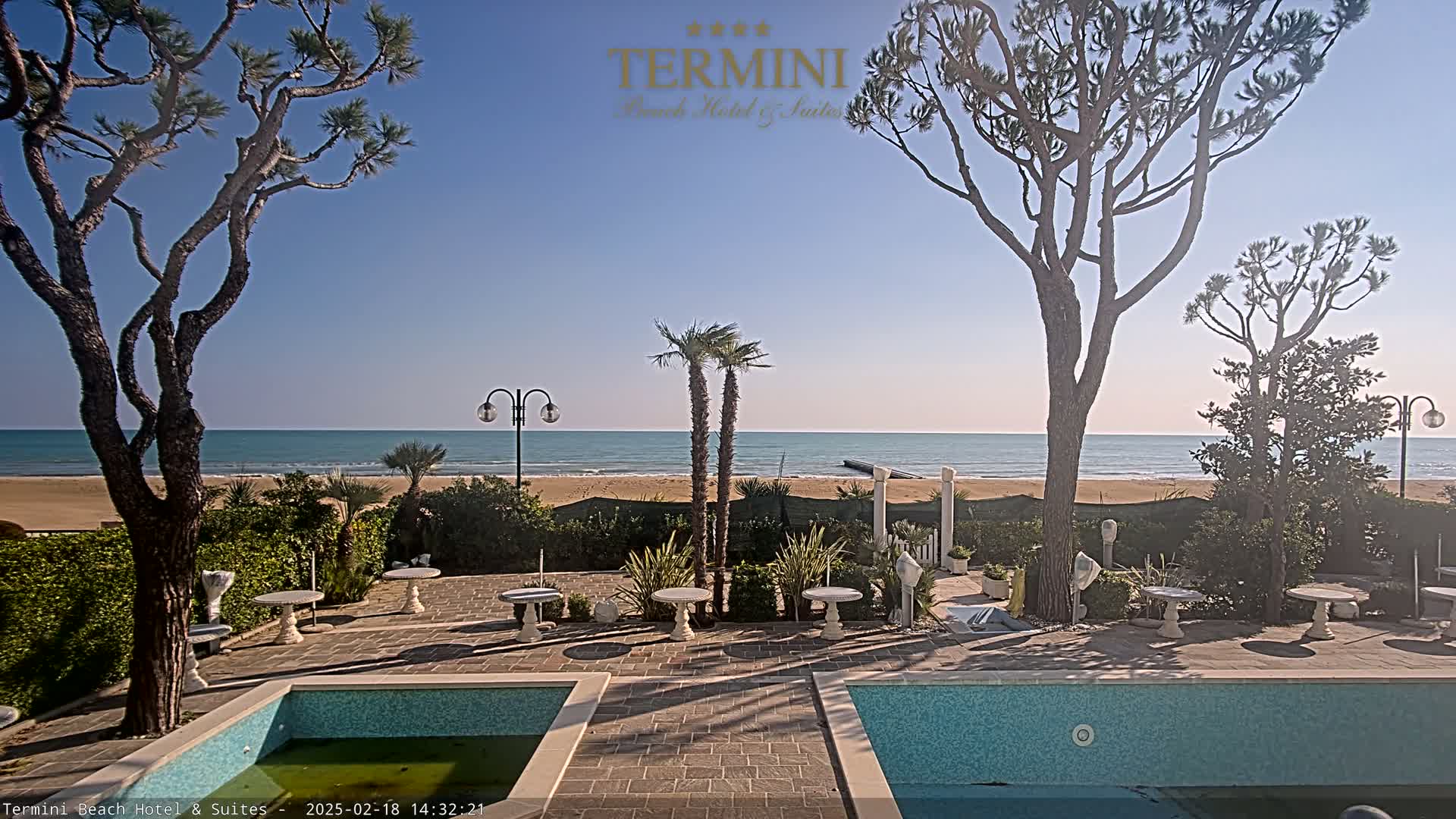 A tranquil seaside patio with two swimming pools, surrounded by palm trees and other vegetation, overlooks a calm ocean under a clear blue sky.