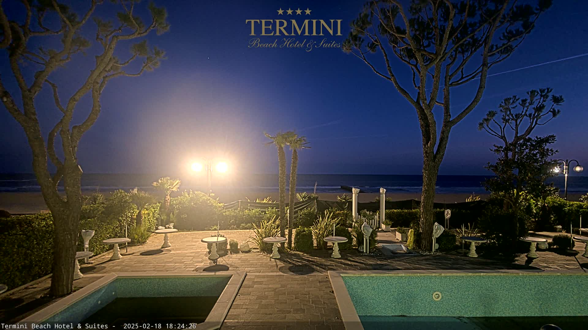 A nighttime view of a seaside patio with two pools, several small tables, and palm trees under a clear, dark blue sky.