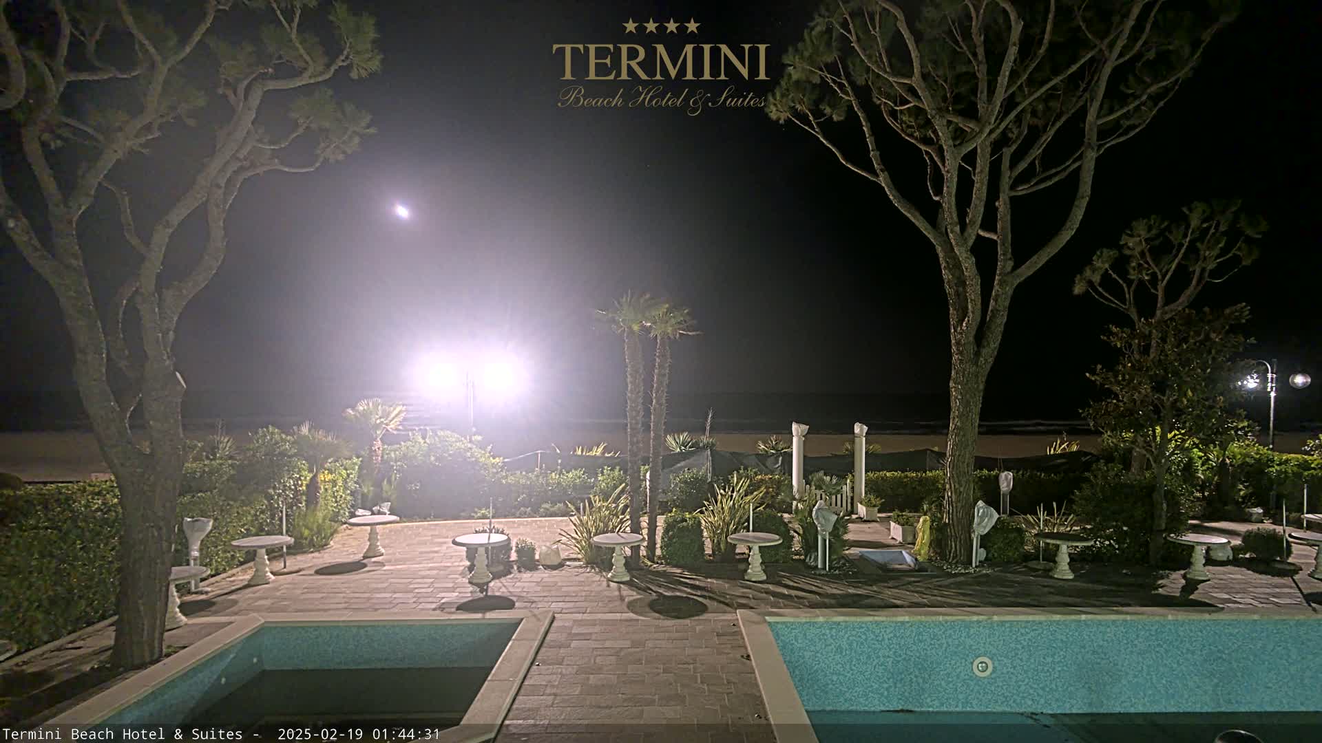 A nighttime view of a hotel patio with a swimming pool and several small tables, situated near a beach under a clear, moonlit sky.