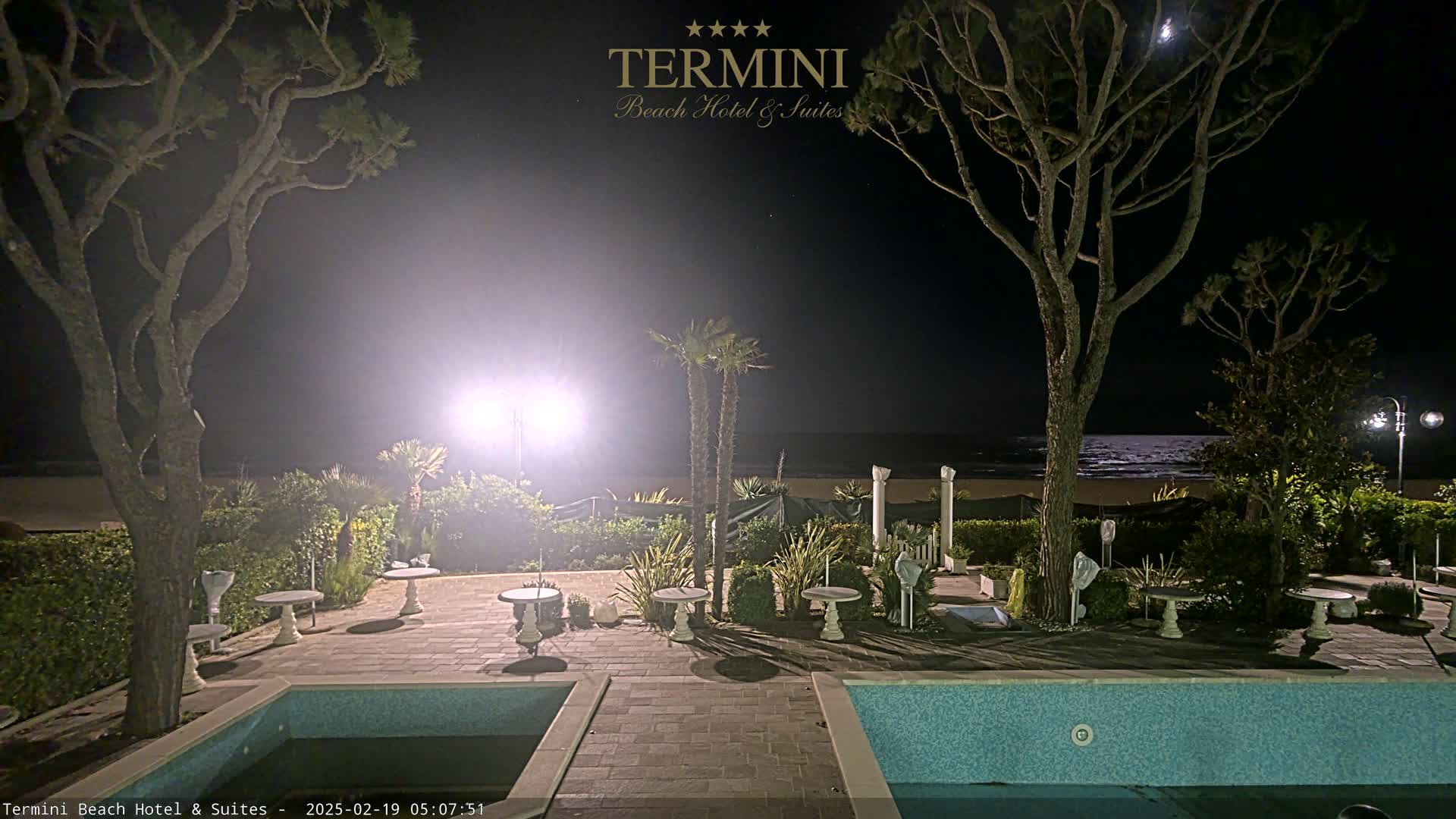 A nighttime view of a patio area with two swimming pools, small tables, and trees, overlooking a calm ocean under a partly moonlit sky.