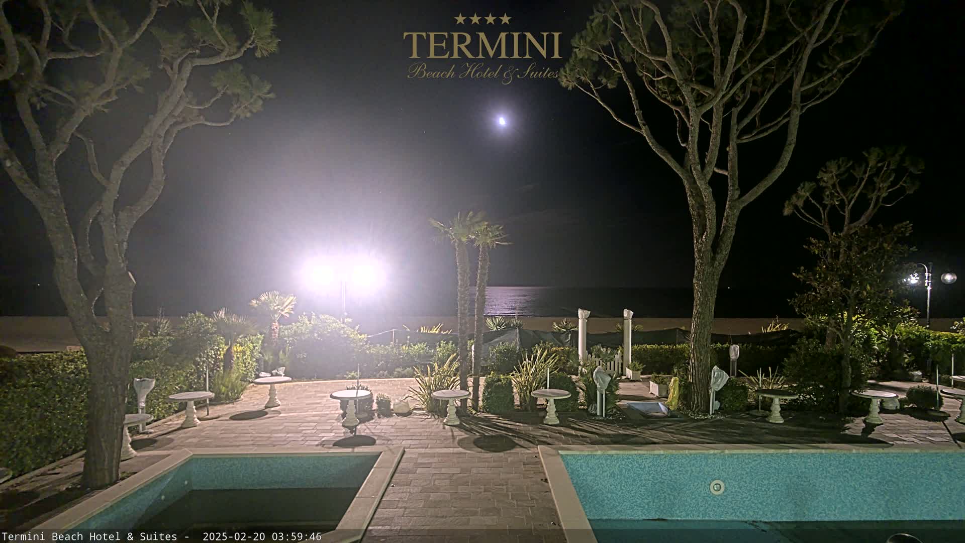 A moonlit night scene shows a patio area with two pools and small tables, surrounded by trees and shrubs, overlooking a calm ocean.