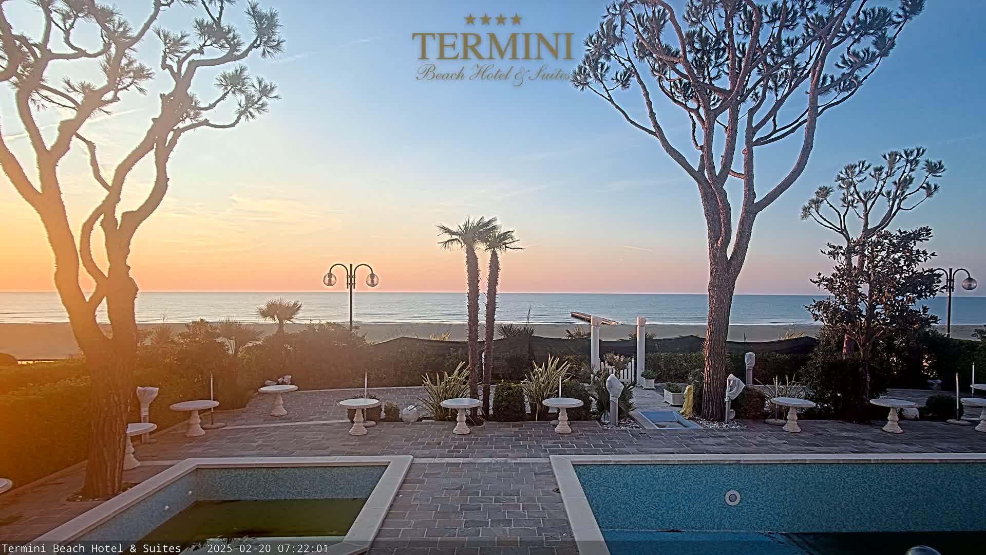 A tranquil beachfront patio at sunrise features two swimming pools, several small tables, and tall, slender trees, with the calm ocean visible in the background.