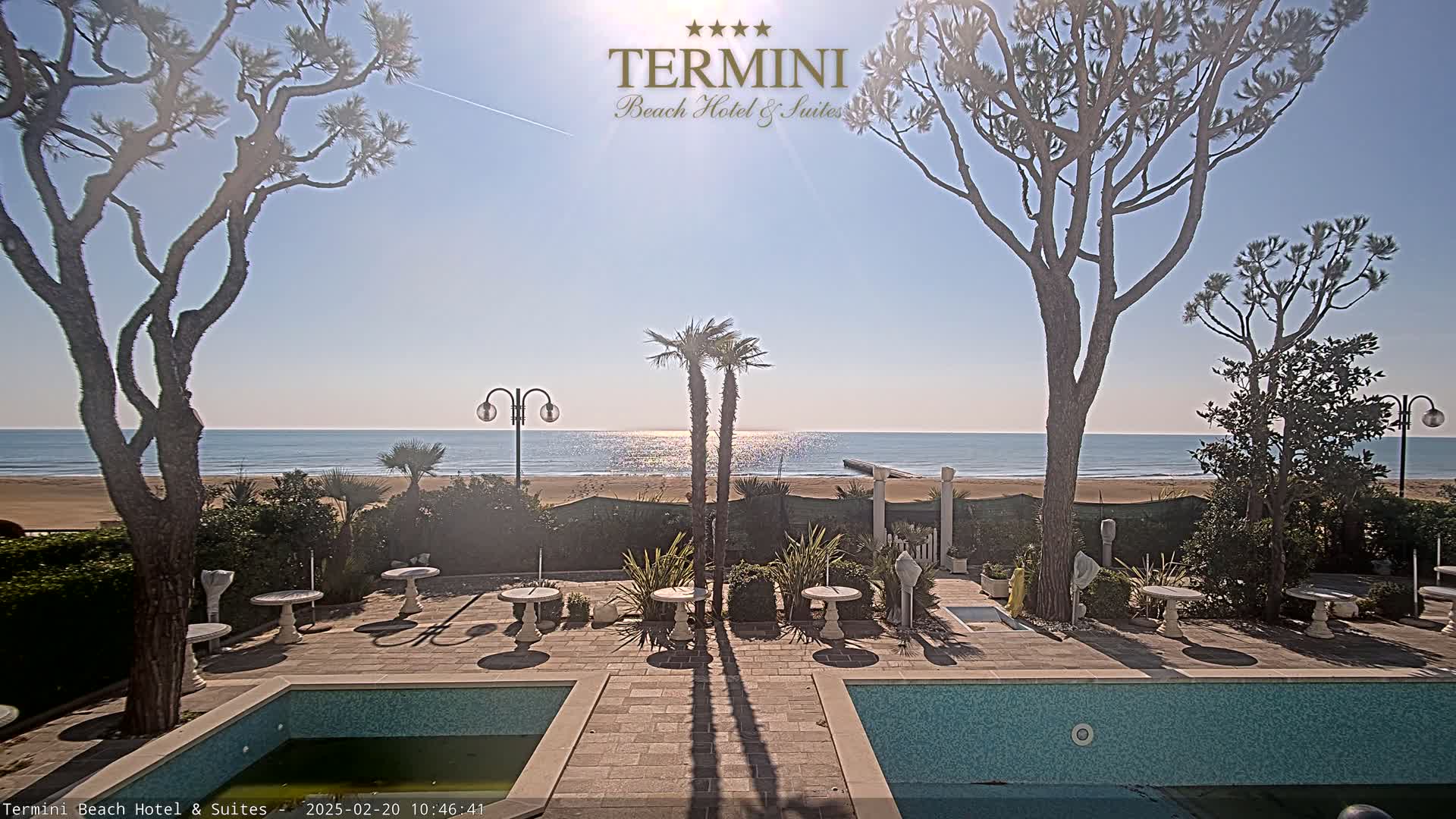 A sunny day at a beachside hotel with a patio containing several small tables, two pools, and palm trees, all overlooking a calm ocean.