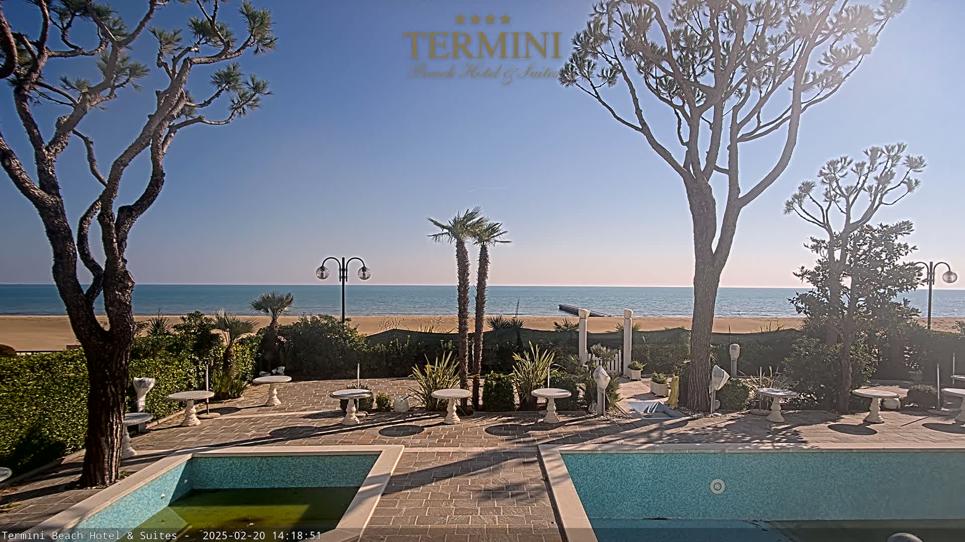 A sunny outdoor patio scene with two swimming pools, palm trees, and a sandy beach with calm ocean in the background.