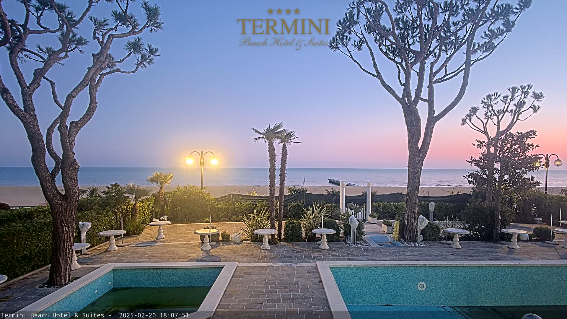 A tranquil outdoor patio scene at twilight shows two swimming pools flanking a paved area with small tables and landscaping, overlooking a calm ocean beach under a pastel sky.