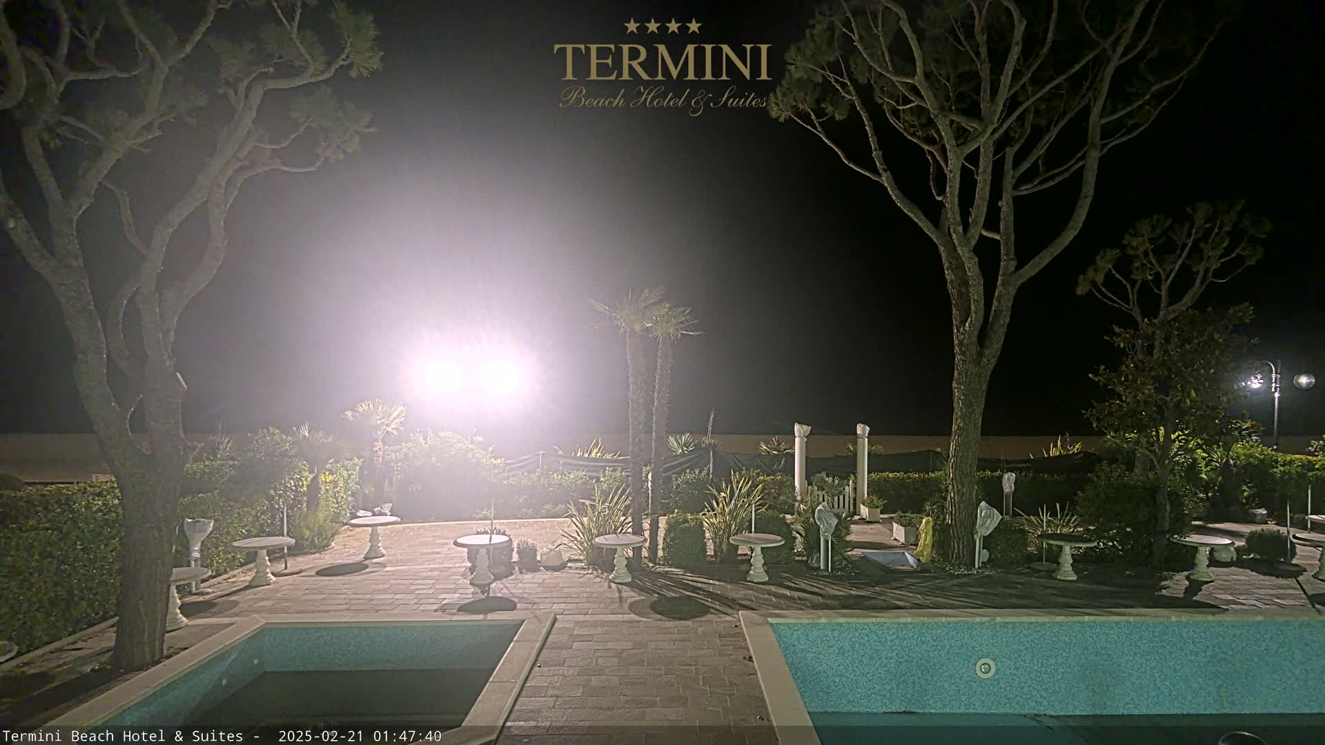 A nighttime view of a patio area with two swimming pools, several small tables, and trees, illuminated by bright lights.