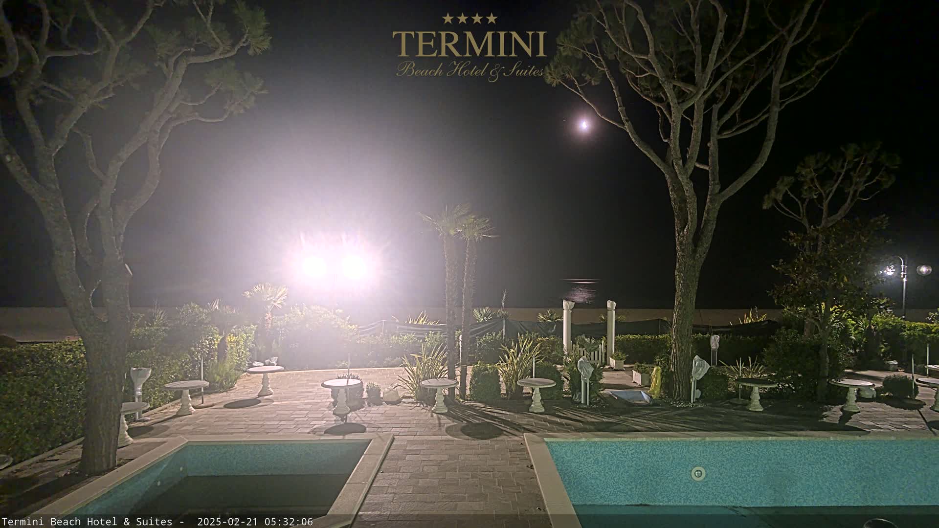 A moonlit night scene shows a patio area with several small tables and chairs surrounding a swimming pool, all situated near a beach under tall trees and bright lights.