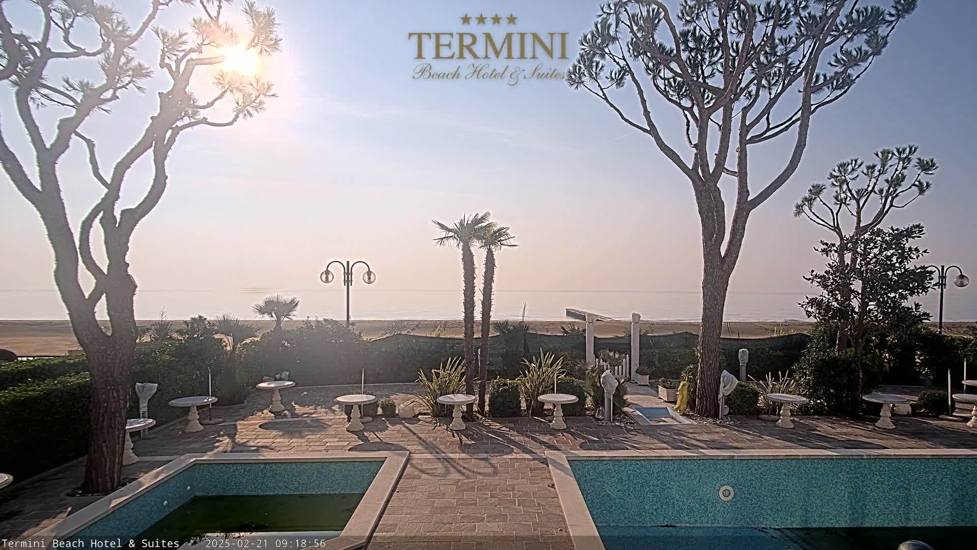 A tranquil outdoor scene shows a patio area with small tables and a swimming pool, overlooking a calm, sandy beach and ocean under a bright, sunny sky.
