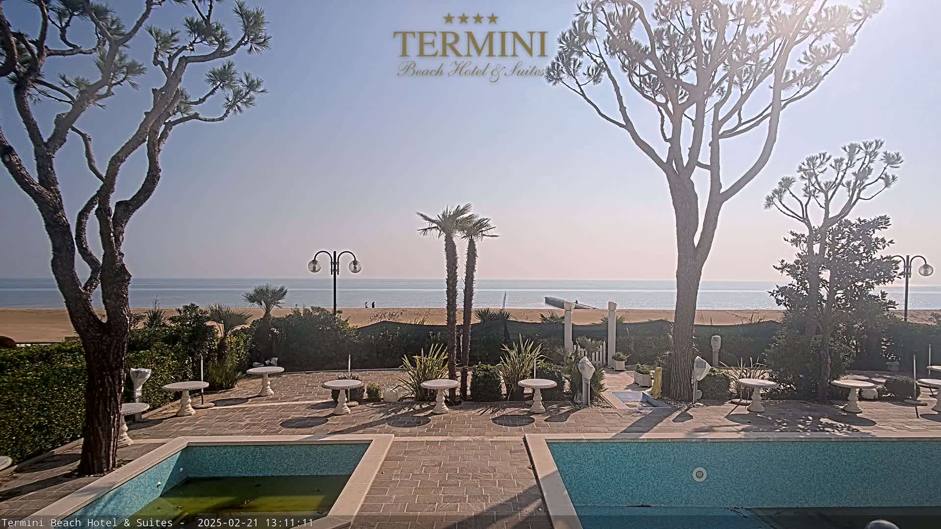 A patio area with two small pools overlooks a sandy beach and calm ocean under a clear, sunny sky.
