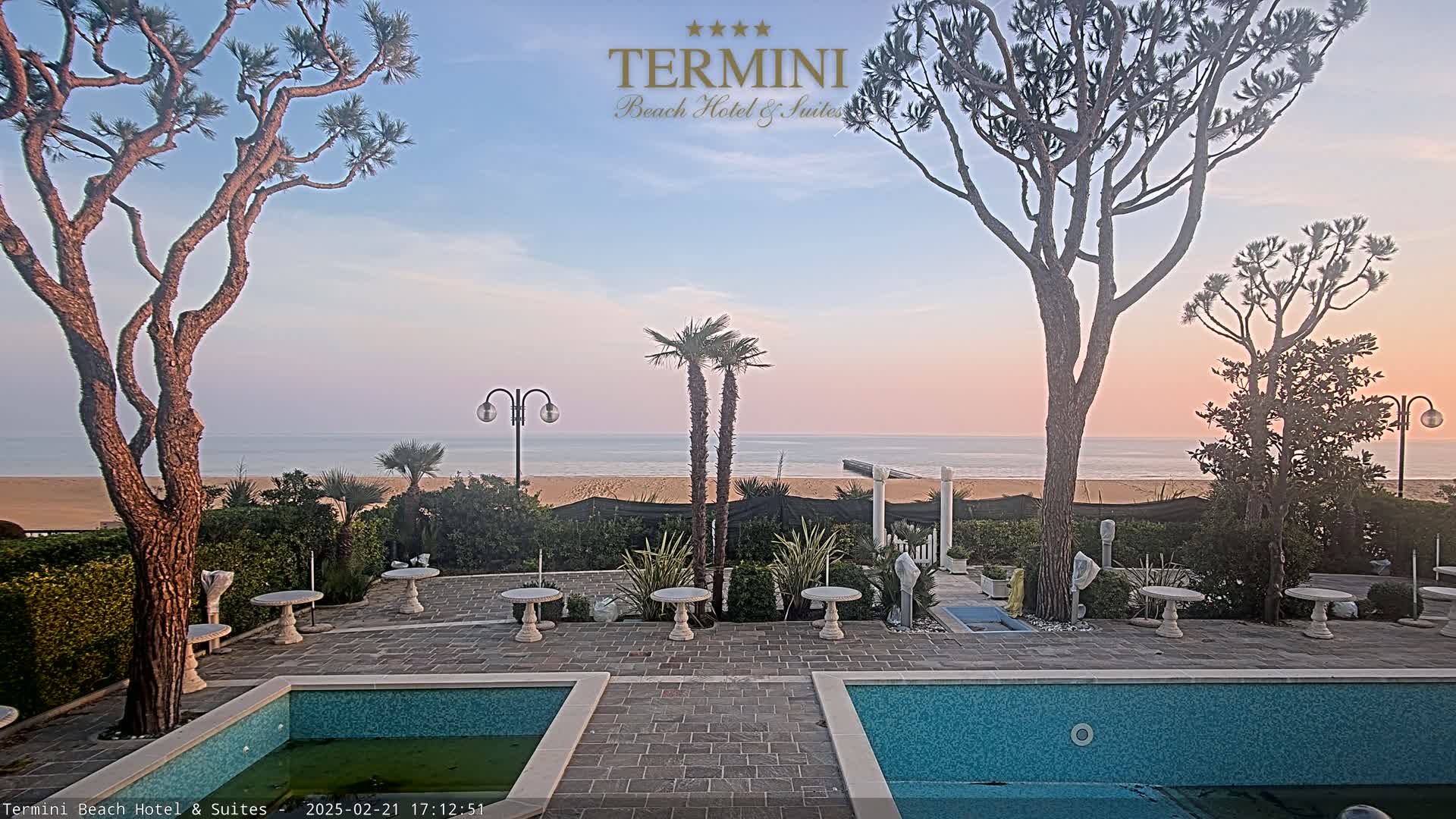 A patio with two pools overlooks a calm ocean beach at sunset.