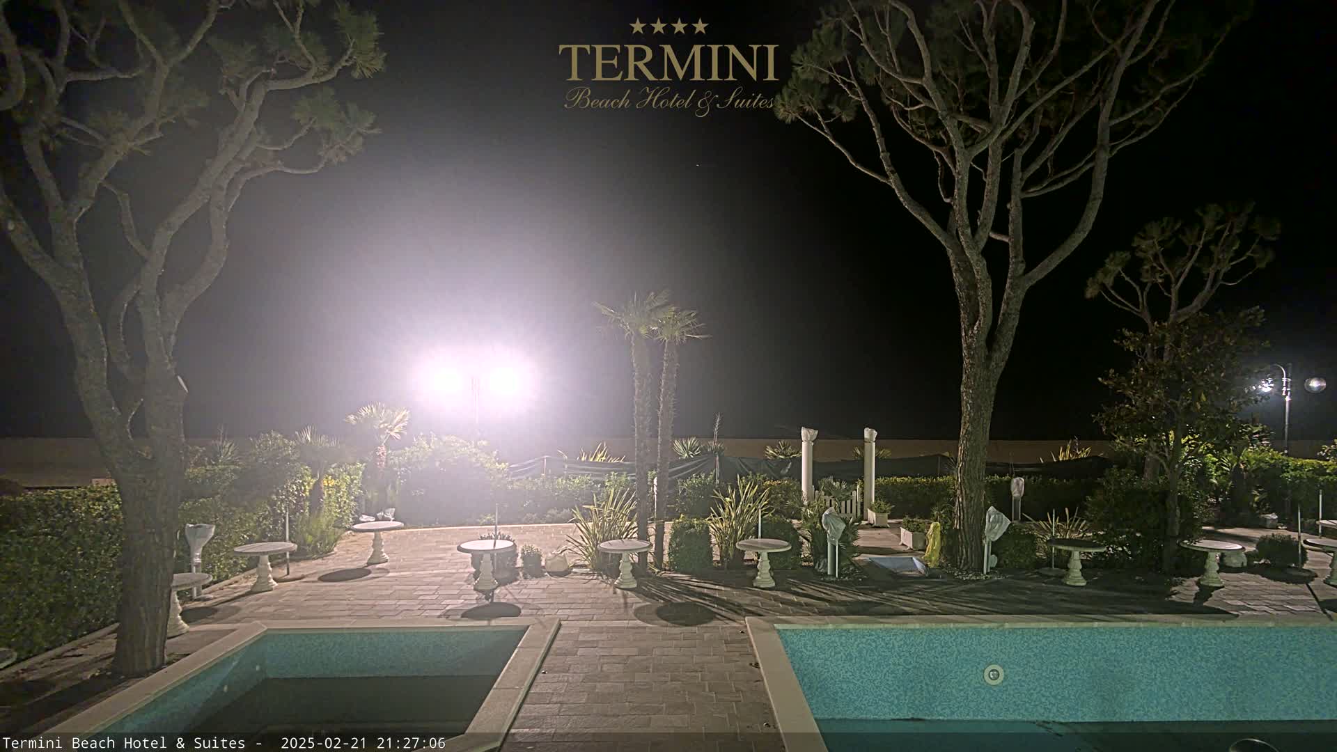 A nighttime view of a patio area with two swimming pools, several small tables and chairs, and trees illuminated by bright lights.
