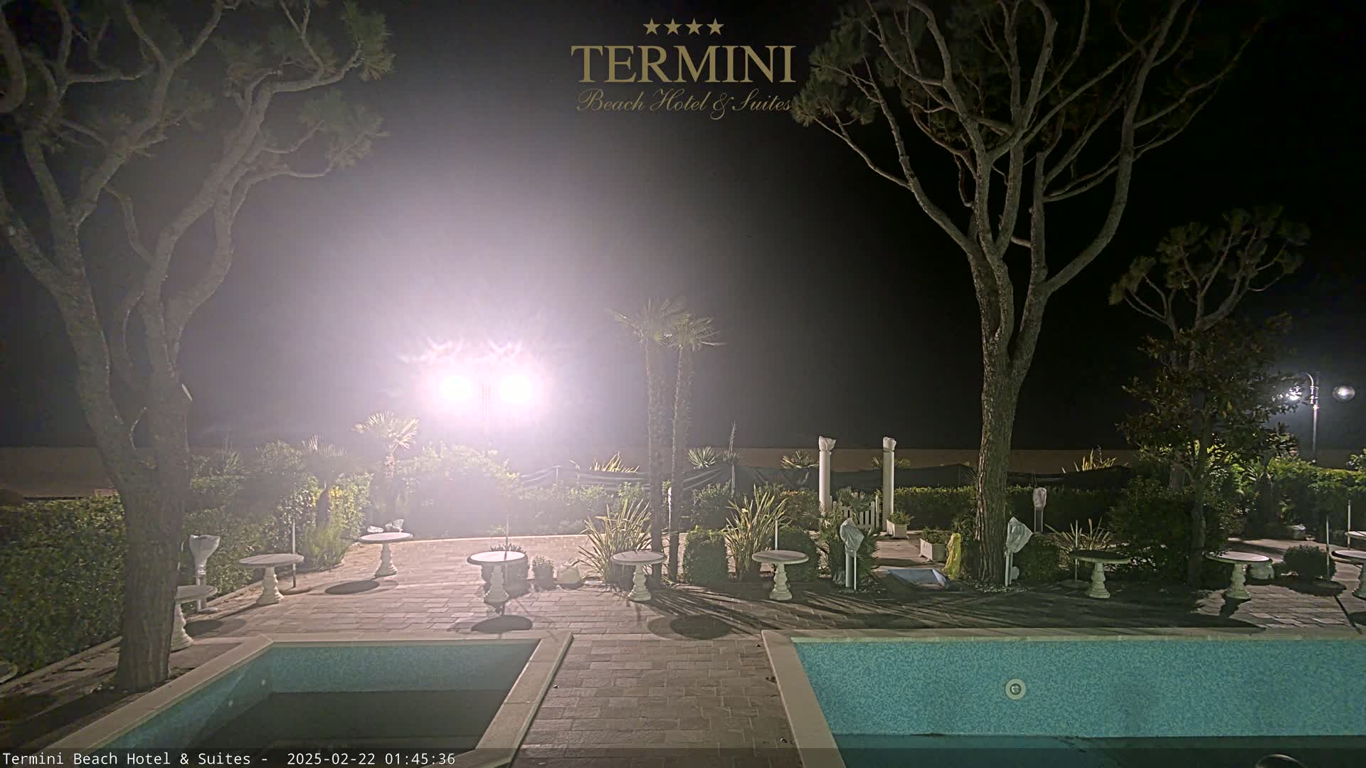 A nighttime view of an outdoor patio area with a swimming pool, several small tables, and trees, illuminated by bright lights.