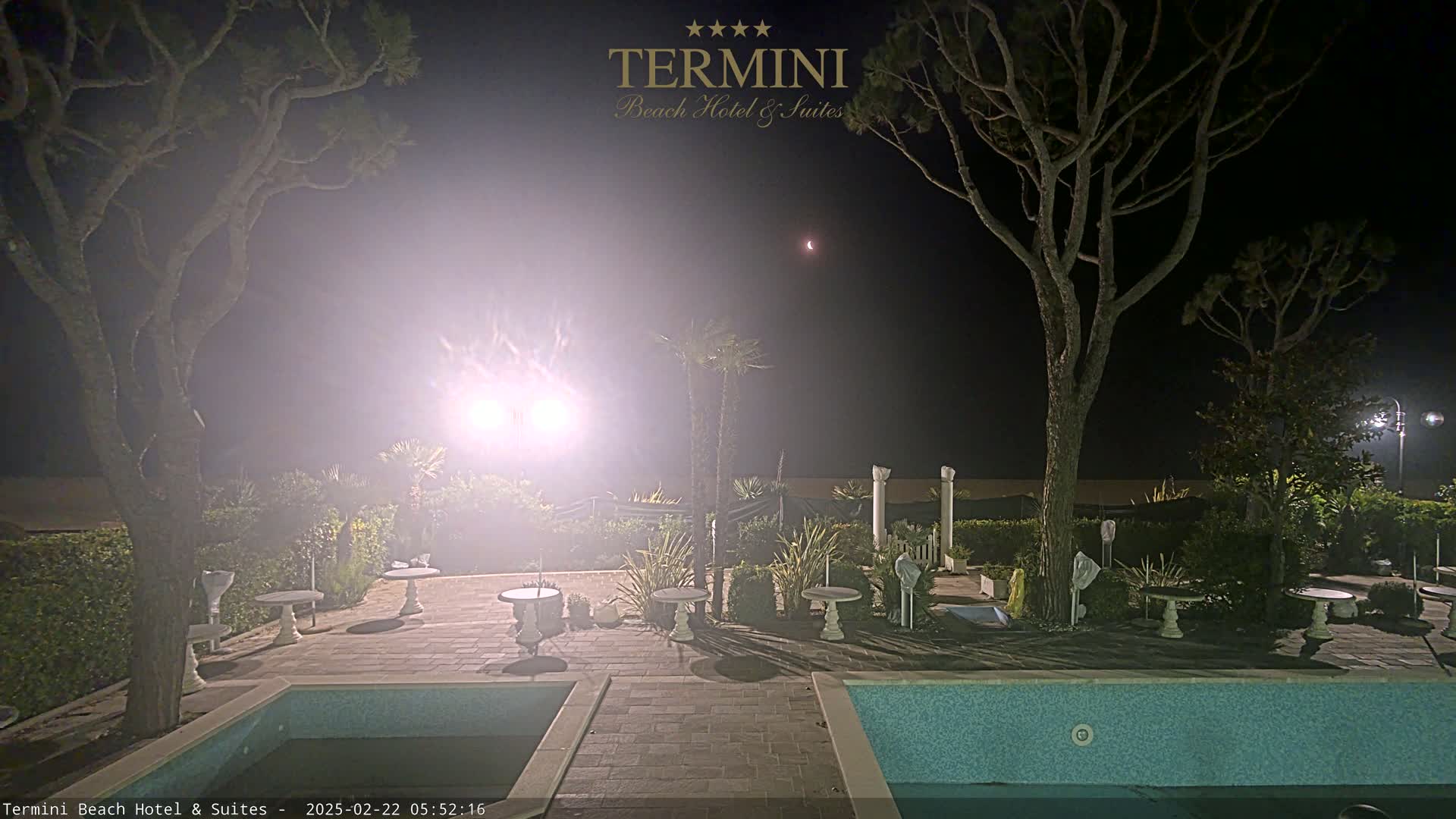 A hazy night scene shows an outdoor pool area with several small tables and chairs surrounding it, illuminated by lights, under a dark sky with a sliver of a moon visible.